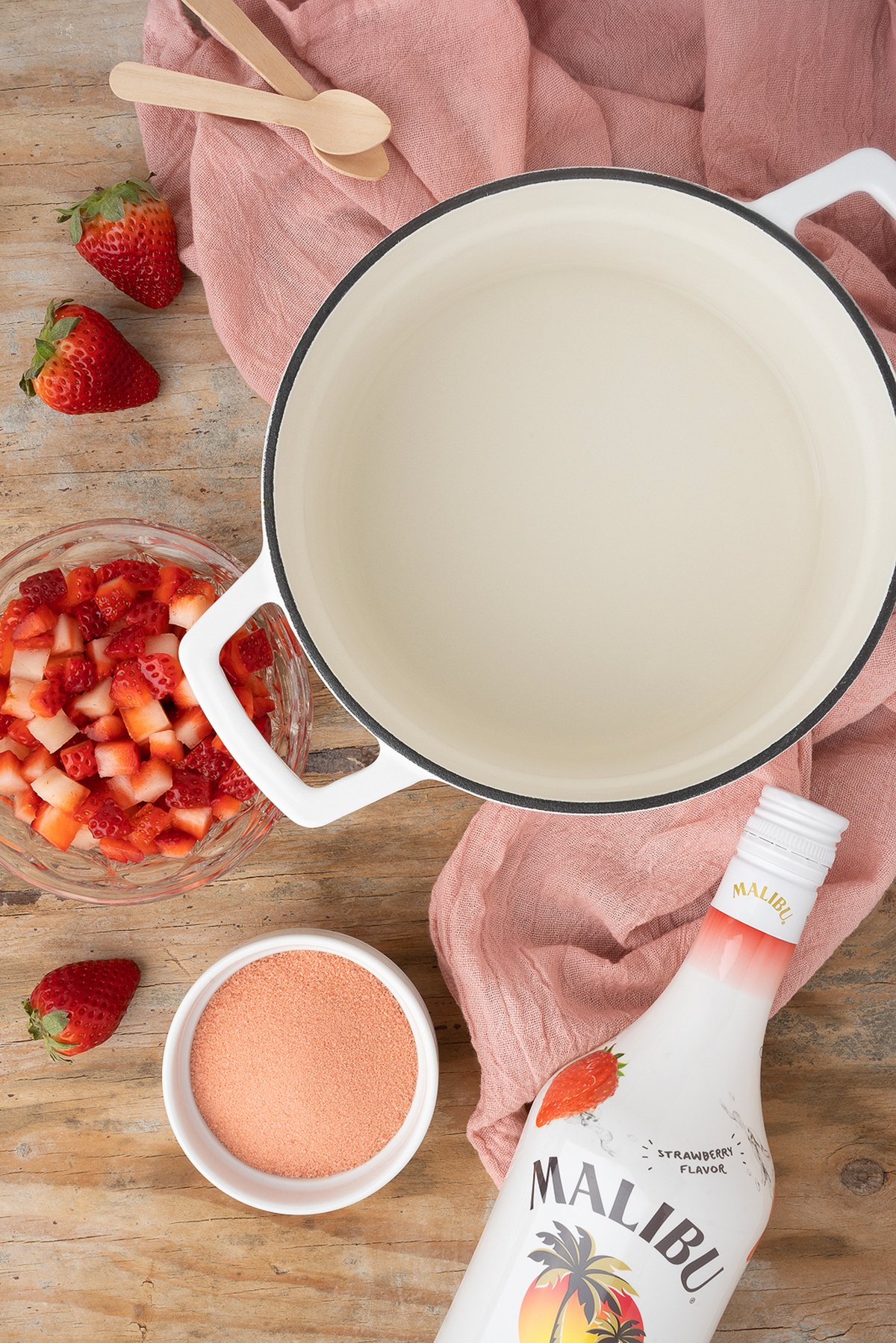 White pot, chopped strawberries, pink powder, Malibu bottle, wooden spoons, and a pink cloth on a table—perfect for whipping up delicious Strawberry Cheesecake Jello Pudding Shots.