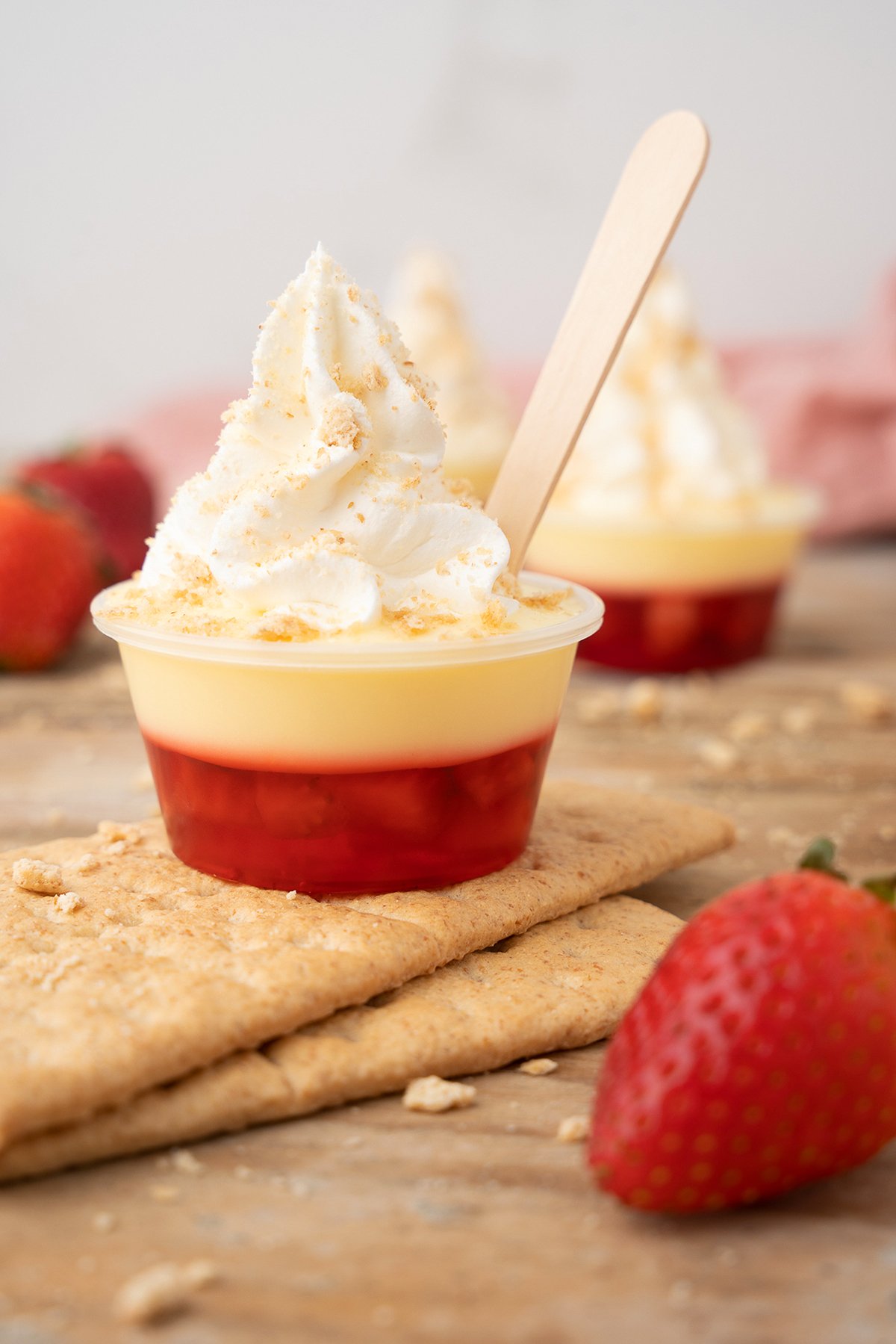 A layered dessert cup with whipped cream, strawberry, and graham crackers, this treat is perfect for fans of strawberry pudding shots, complete with a wooden spoon on top.