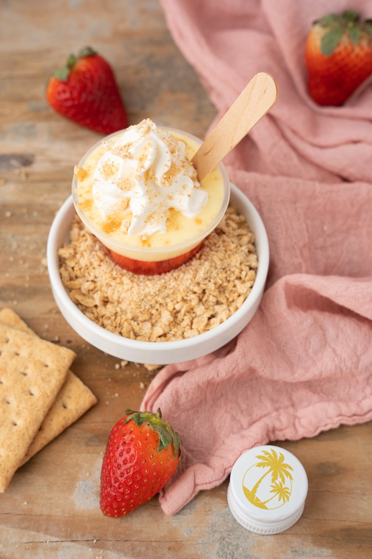 A cup of pudding with whipped cream and a wooden spoon, surrounded by strawberries, graham crackers, and crumbs—perfect for making strawberry pudding shots or indulgent cheesecake shooters.