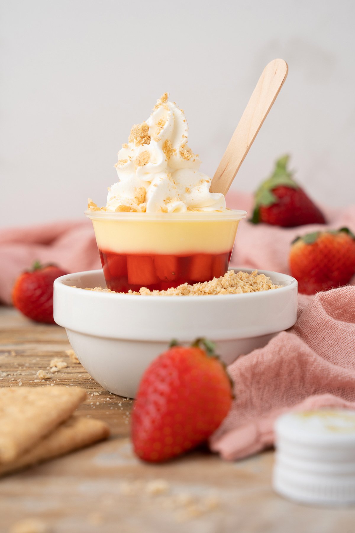 A layered dessert cup with whipped cream, a wooden spoon, and fresh strawberries on a wooden table, perfect for enjoying as strawberry pudding shots or a delightful strawberry dessert cocktail.