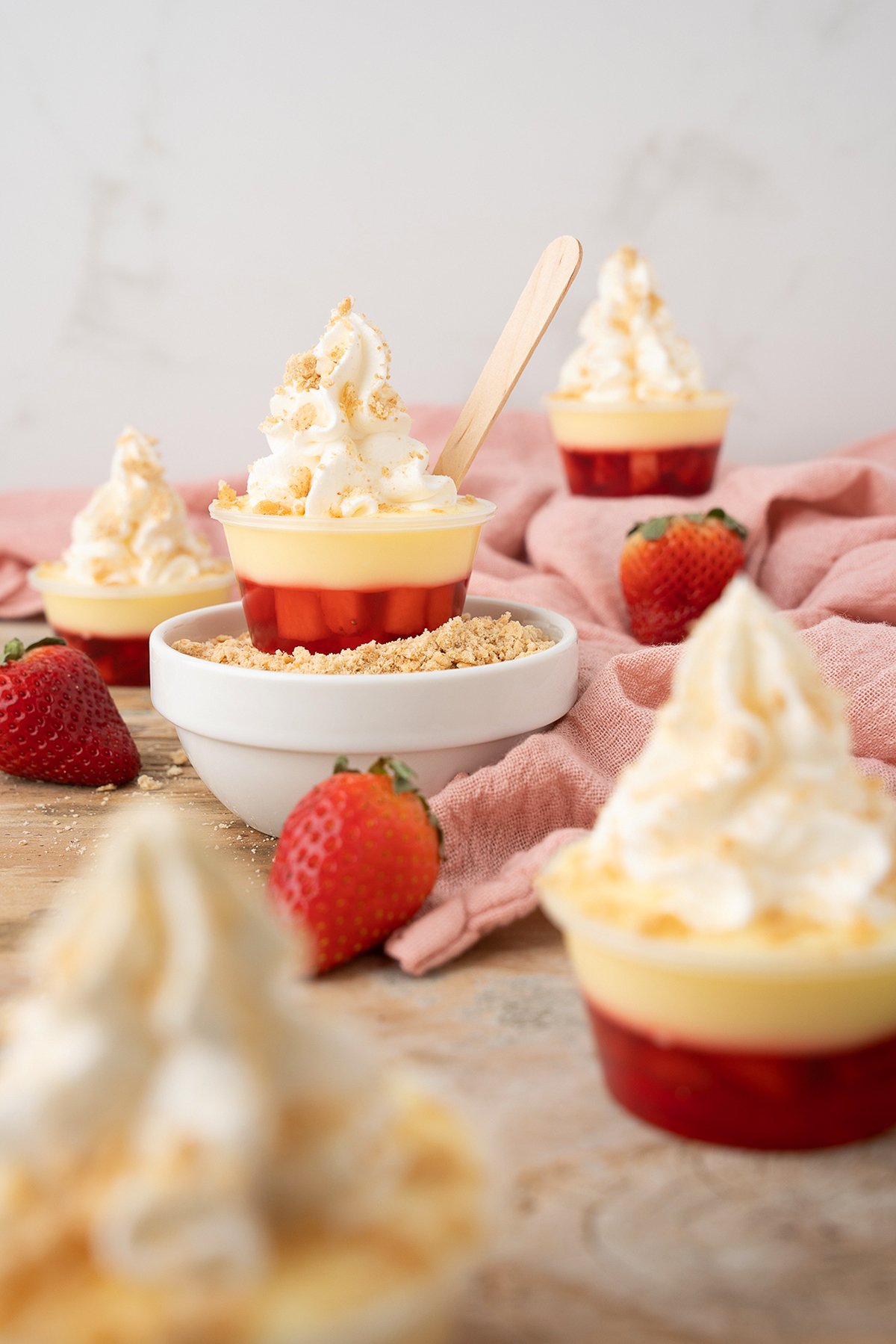 Small cups of layered cheesecake shooters with whipped cream, strawberries, and crumbs sit on a rustic table draped with a pink cloth.