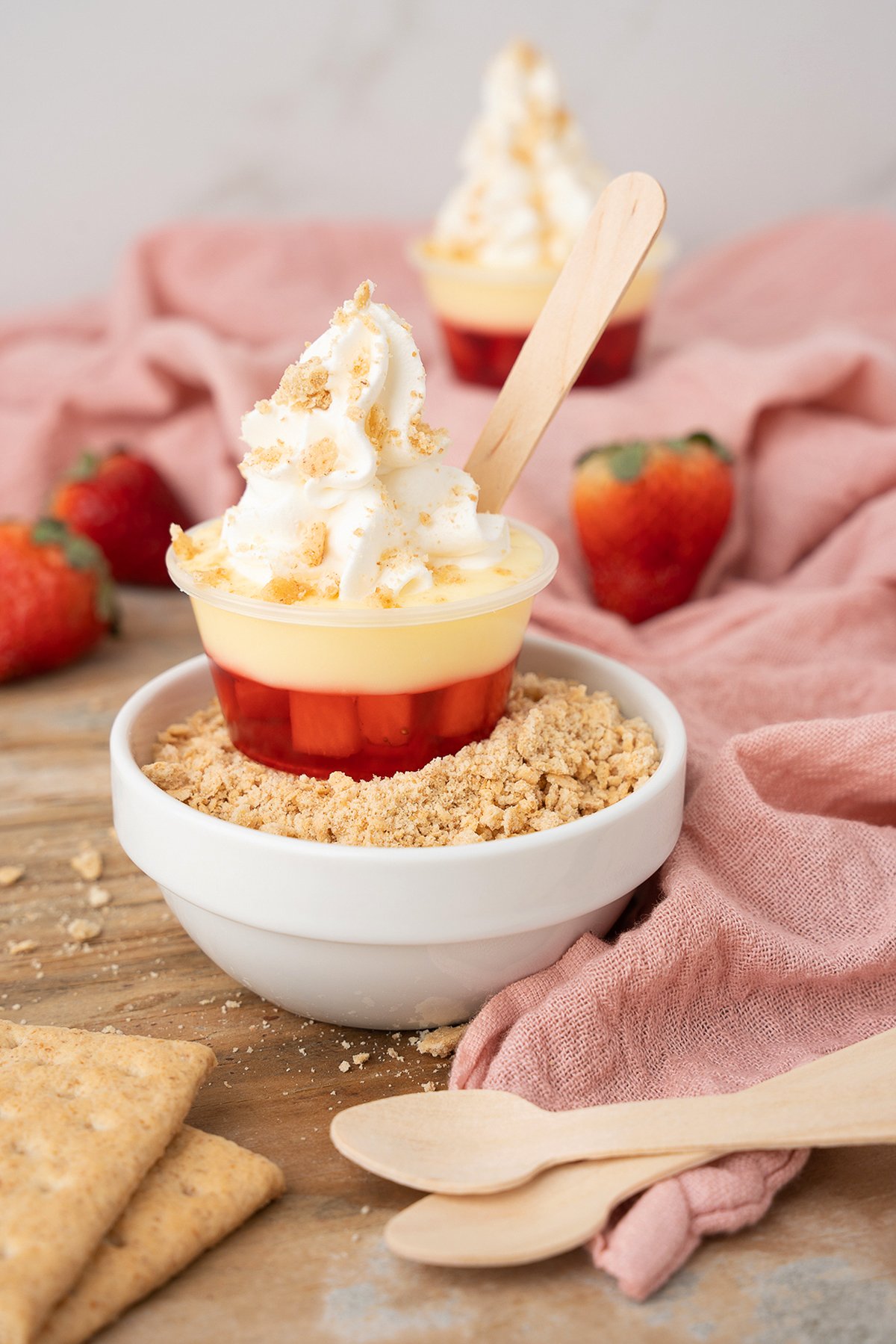 Mini strawberry parfait with whipped cream in a cup, set in a bowl of graham cracker crumbs, with strawberries—perfect as cheesecake shooters for a delightful treat.