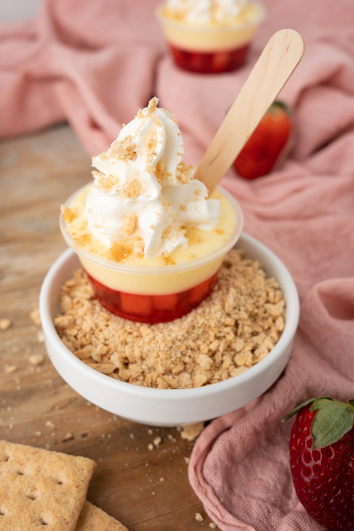 Mini strawberry parfait cup with whipped cream, set in a bowl of crushed graham crackers, just like a bite-sized Strawberry Cheesecake Jello Pudding Shot, served with a wooden spoon.