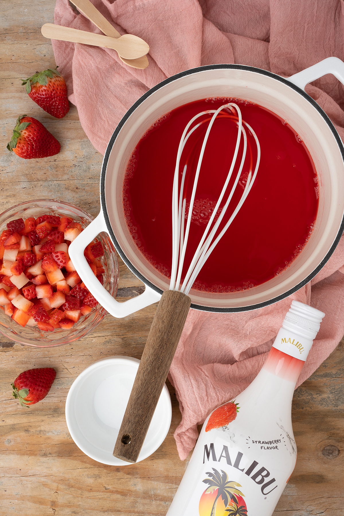 A pot of red liquid with a whisk, diced strawberries, Malibu bottle, and pink cloth on a wooden table—perfect ingredients for Strawberry Cheesecake Jello Pudding Shots.