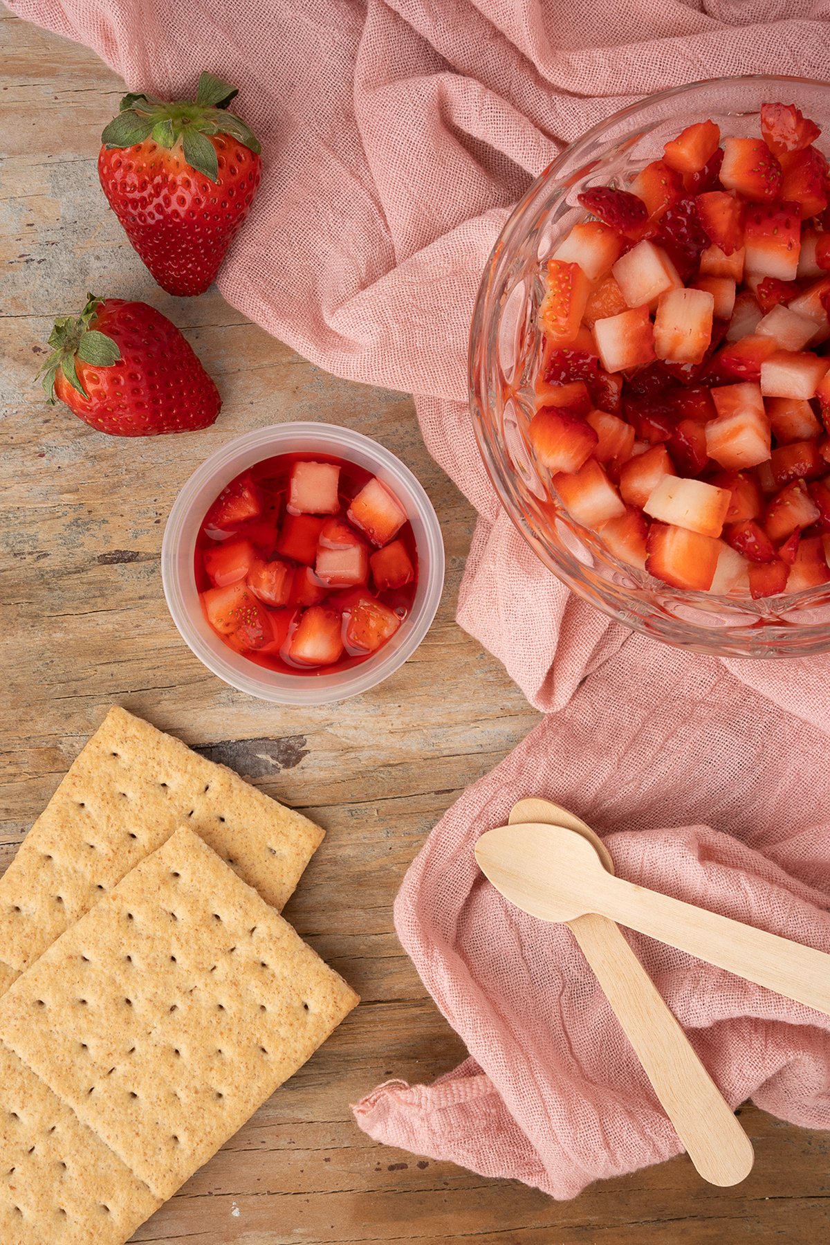 Diced strawberries in bowls, whole strawberries, graham crackers, and wooden spoons on a pink cloth—perfect toppings for Strawberry Cheesecake Jello Pudding Shots.