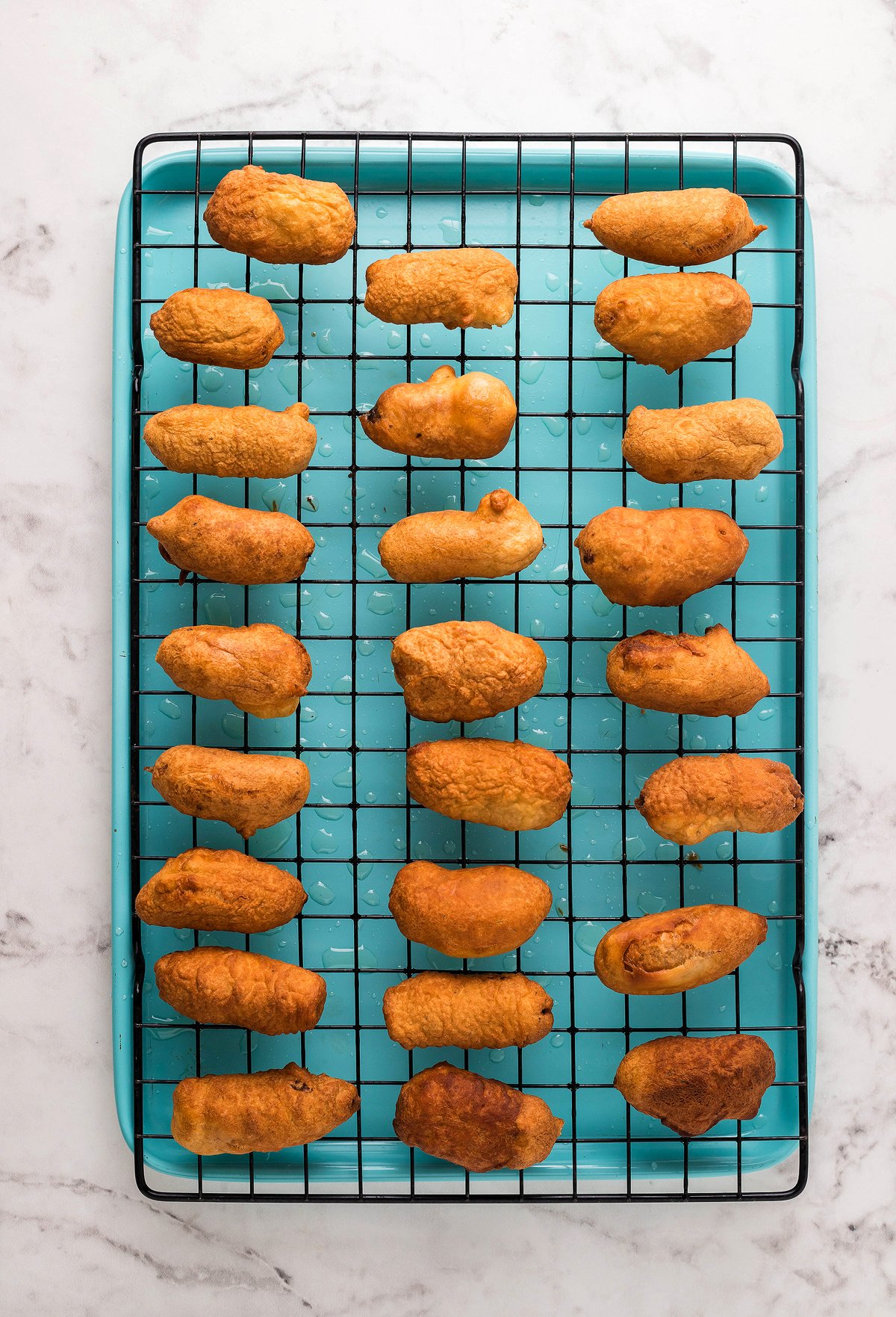 Rows of golden-brown beer battered little smokies cooling on a wire rack over a blue tray—perfect as a game day appy on your marble countertop.