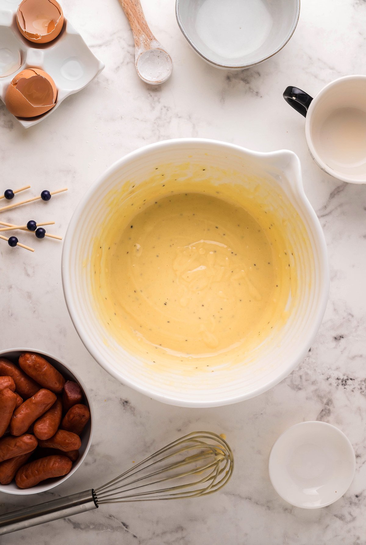 A mixing bowl of yellow batter surrounded by sausages, a whisk, and ingredients on a marble countertop—perfect for whipping up an appetizer or finger food for your next game day appy.