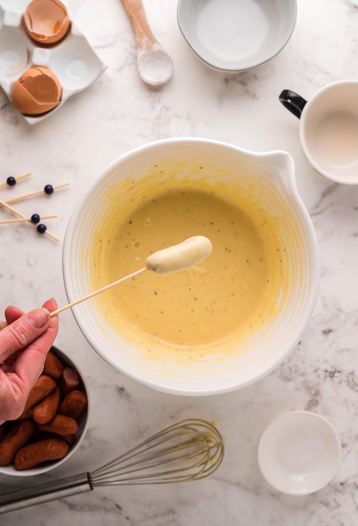A hand dips a cocktail weenie on a skewer into a bowl of thick yellow batter on a marble countertop, perfect for beer battered little smokies.