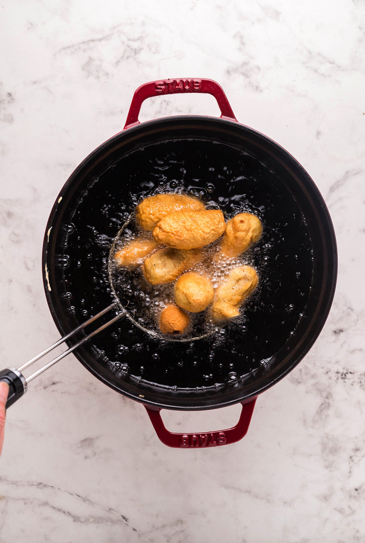 Golden brown dough balls frying in hot oil inside a red pot, with a hand holding a slotted spoon—perfect as an appetizer or finger food. Serve them alongside easy horseradish mayo dipping sauce for an extra kick.