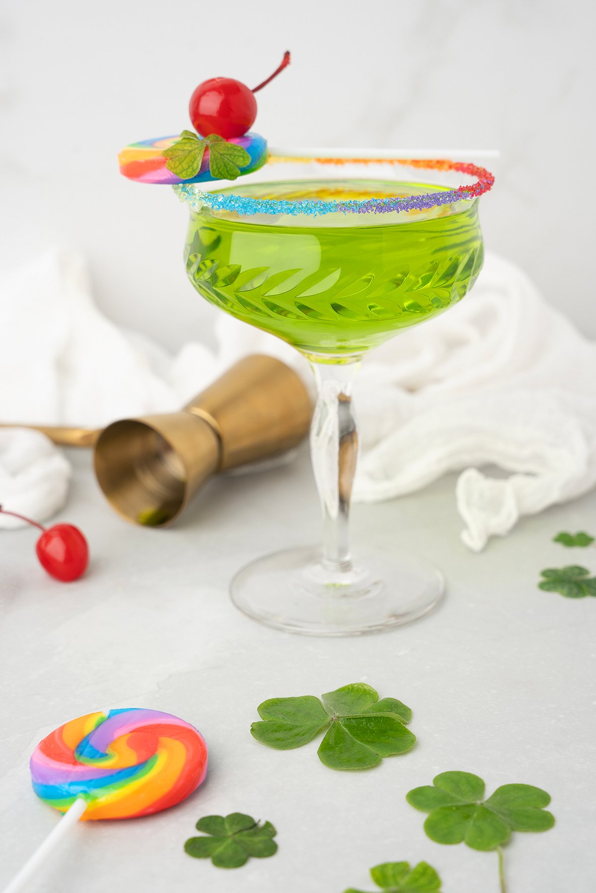 A green cocktail made with Irish whiskey and melon liqueur sits in a glass with a rainbow rim, topped with a cherry and lollipop, surrounded by clovers.