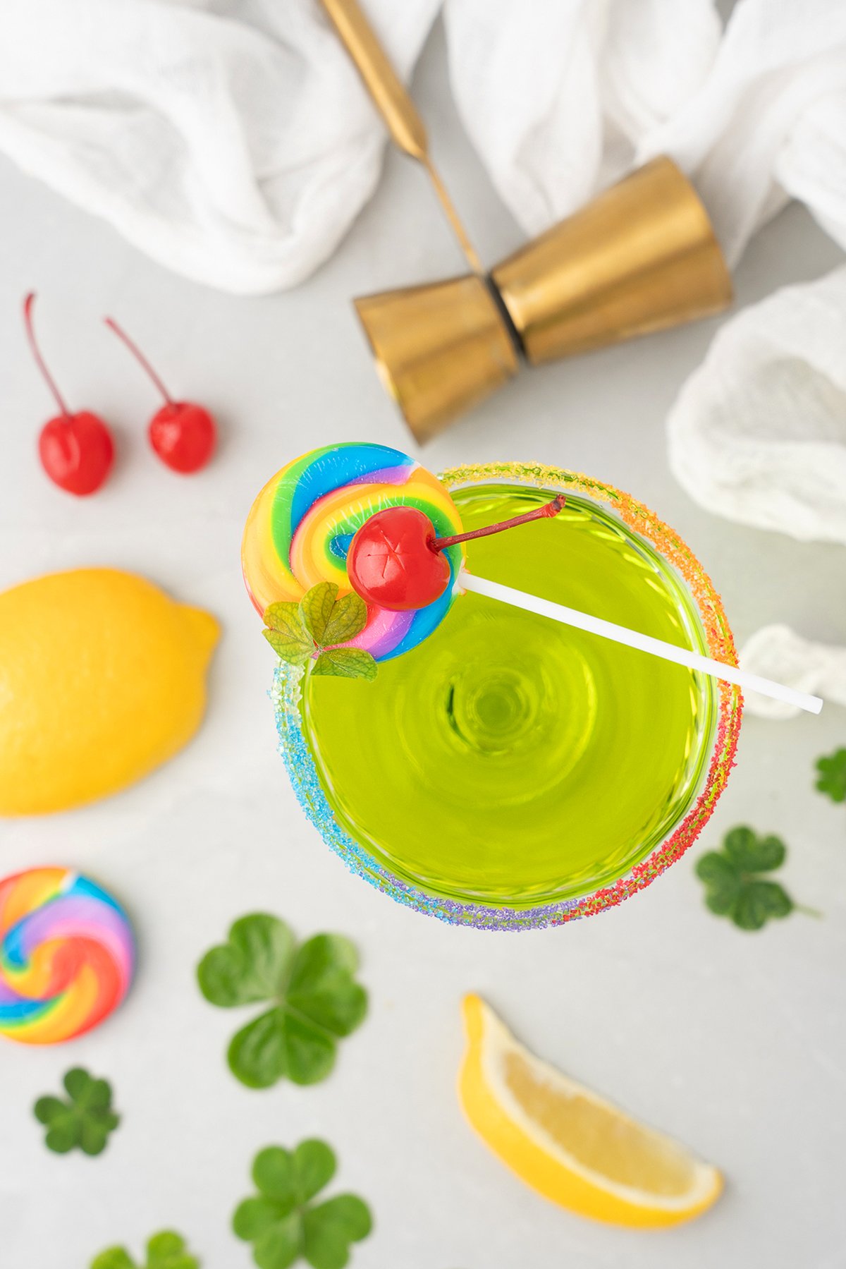 A green Rainbow Irish Whiskey Cocktail with a rainbow sugar rim, cherry and lollipop garnish, surrounded by lemon wedges and clovers.