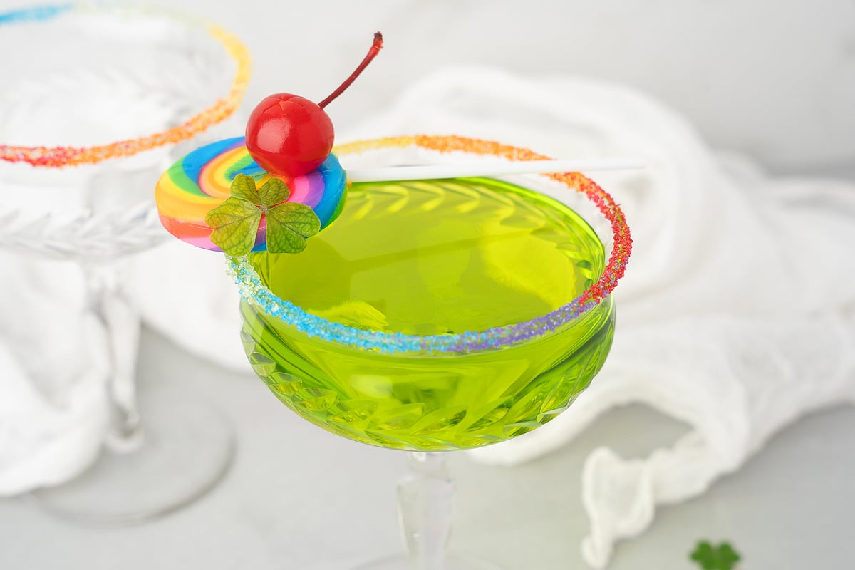 A green Rainbow Irish Whiskey Cocktail in a glass with a rainbow sugar rim, made with sour apple schnapps, topped with a cherry and a rainbow lollipop.