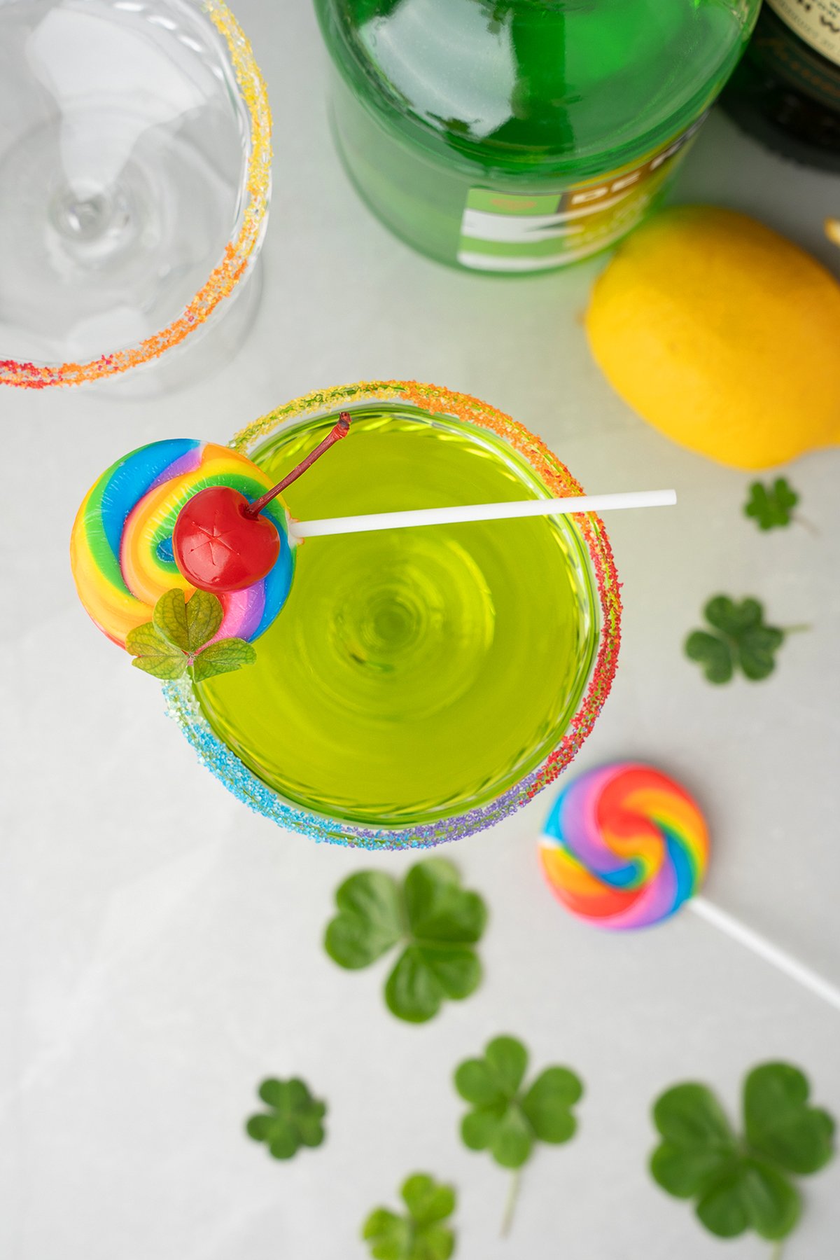 Green drink with rainbow sugar rim, a cherry, shamrock, and rainbow lollipop garnish; made with Irish whiskey and melon liqueur—perfect for St. Patrick’s Day. Lemon and clovers sit nearby.