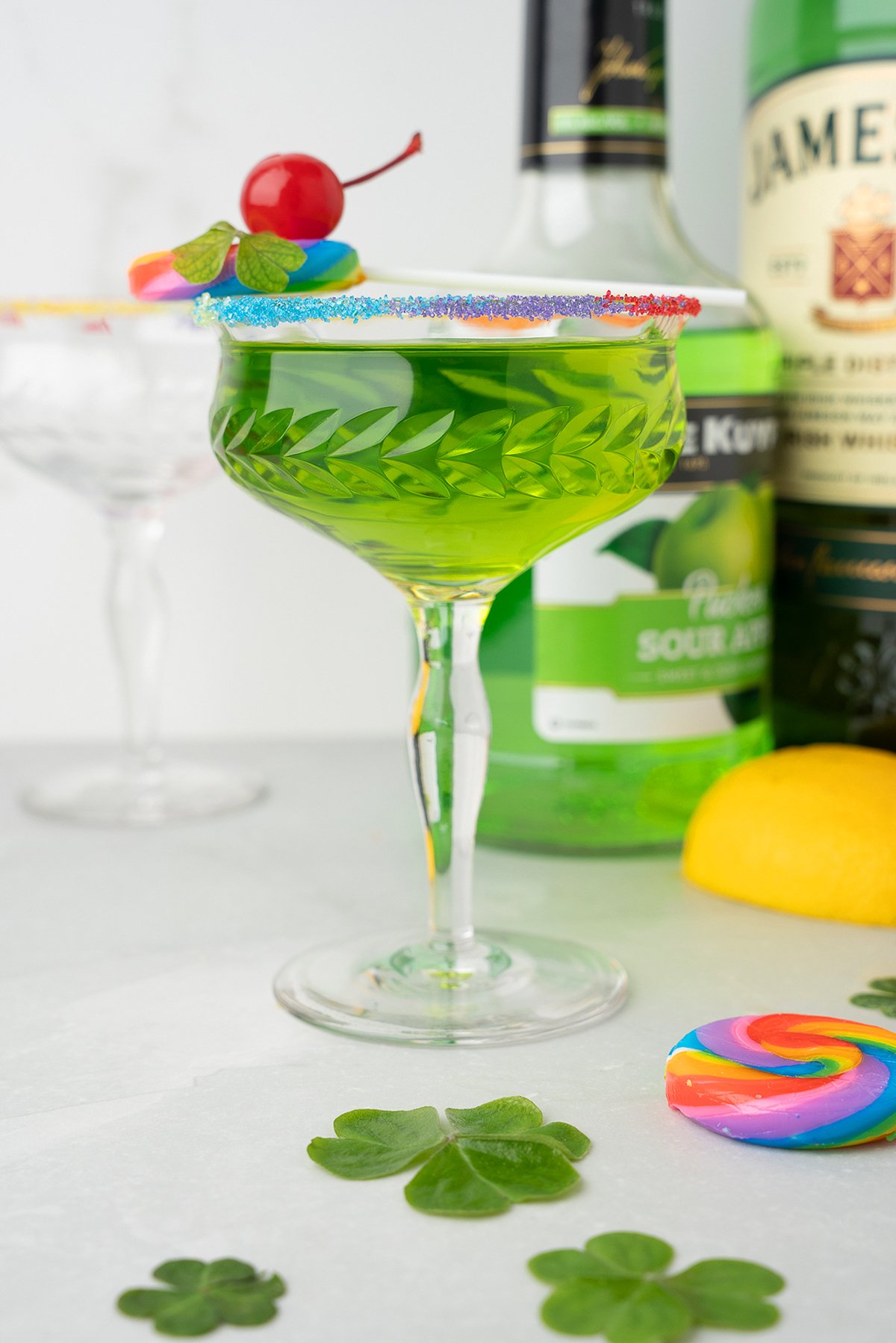 A green Rainbow Irish Whiskey Cocktail in a glass with a rainbow sugar rim, cherry, mint, and lollipop, next to bottles and a lemon, made with Irish whiskey for a vibrant twist.