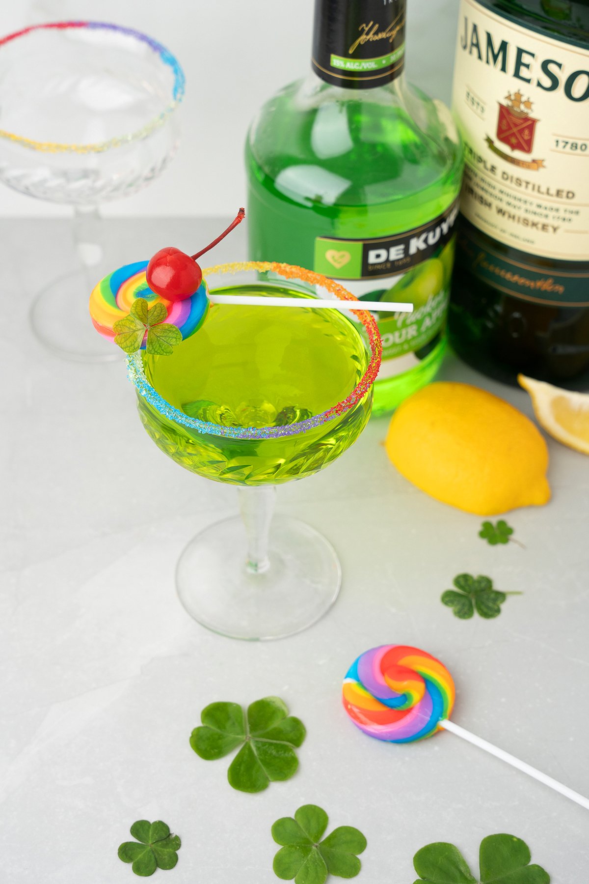 A green cocktail made with Irish whiskey and melon liqueur, topped with a rainbow lollipop, cherry, and colorful rim—surrounded by clovers, lemon, and bottles.