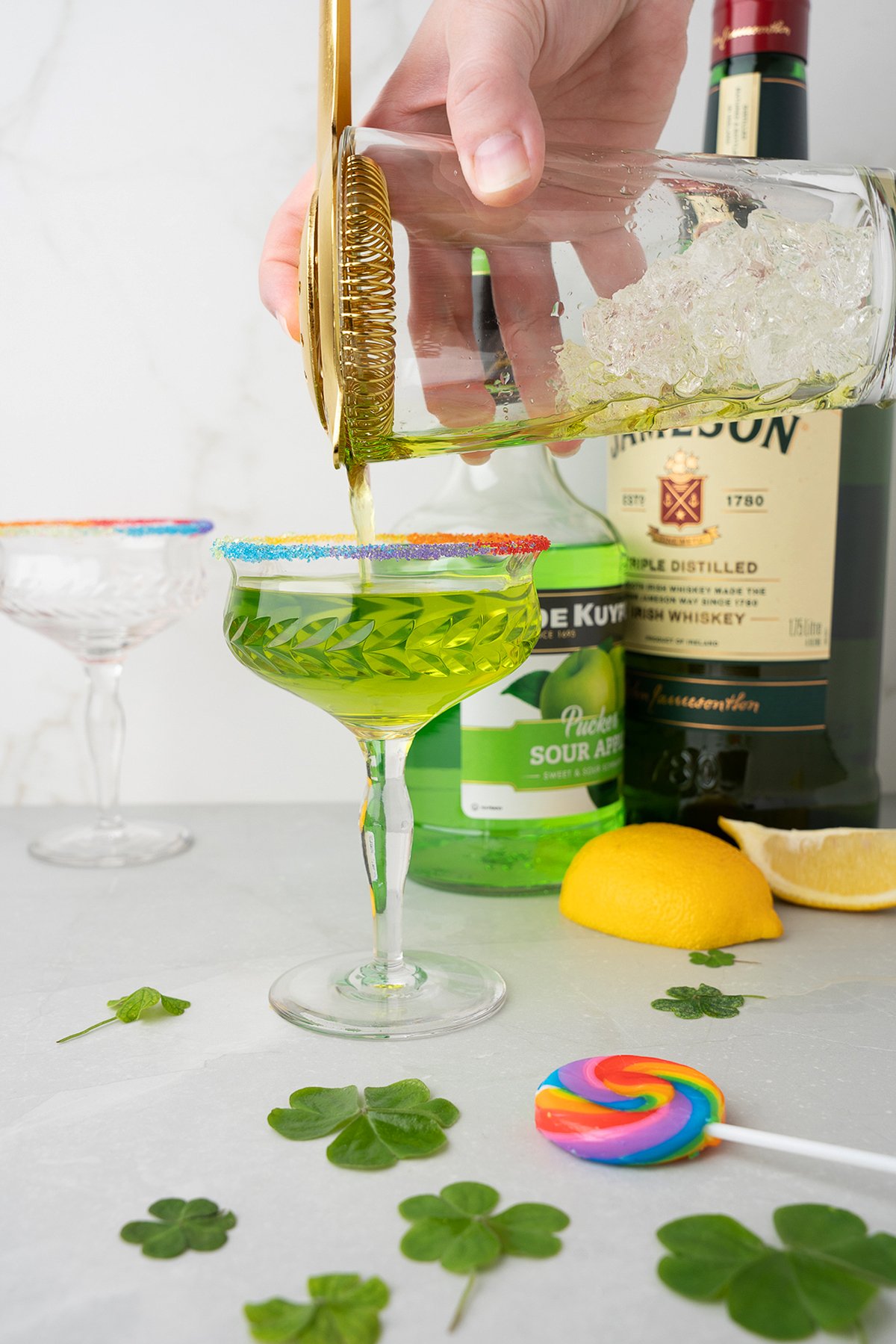 A green Rainbow Irish Whiskey Cocktail is being poured into a glass with a rainbow sugar rim, surrounded by shamrocks and lemons, featuring the tangy flavors of sour apple schnapps and melon liqueur.
