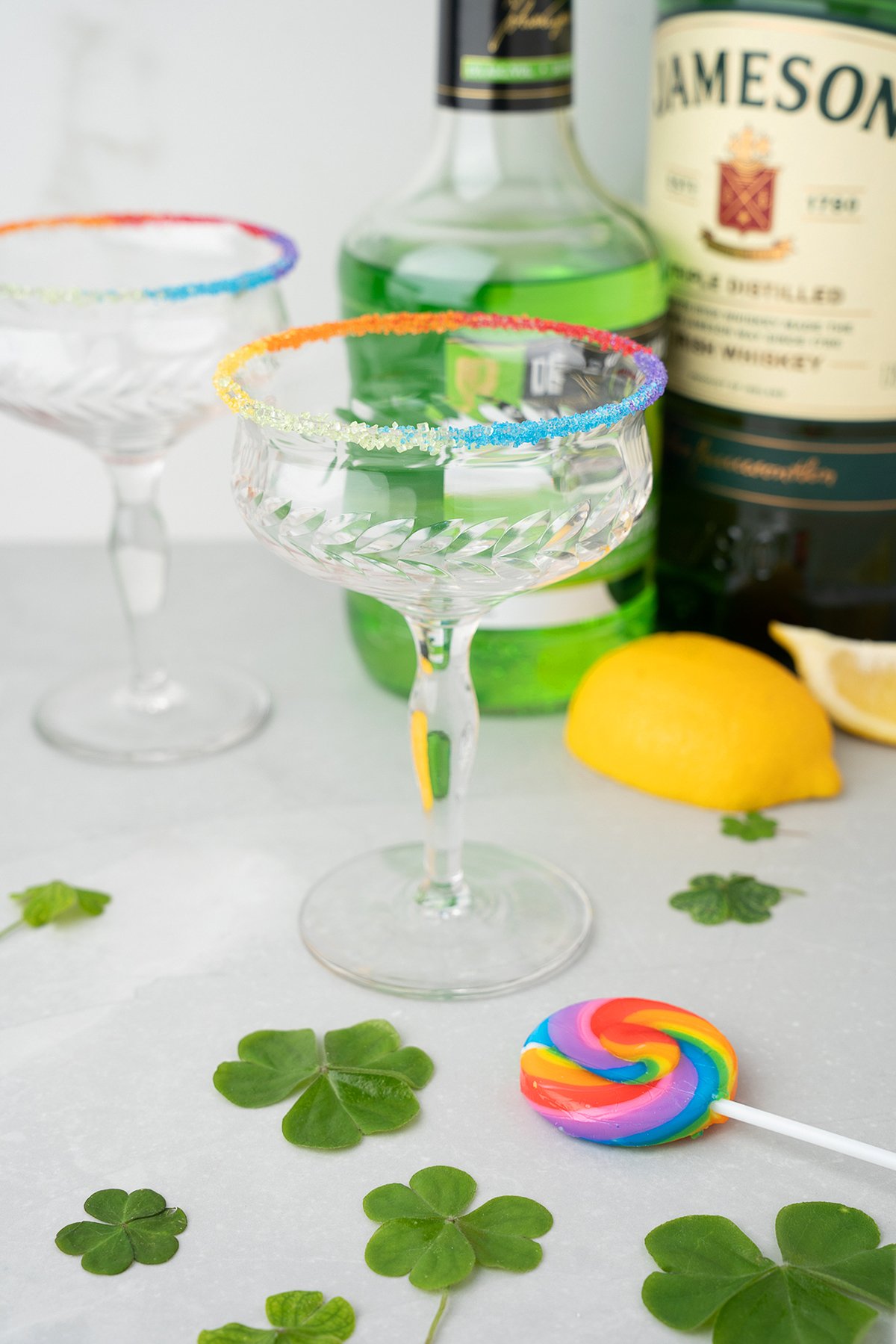 Two glasses with a rainbow sugar rim, bottles of melon liqueur, a lemon, shamrock leaves, and a rainbow lollipop decorate the table—perfect for celebrating St. Patrick’s Day or Pride Day.