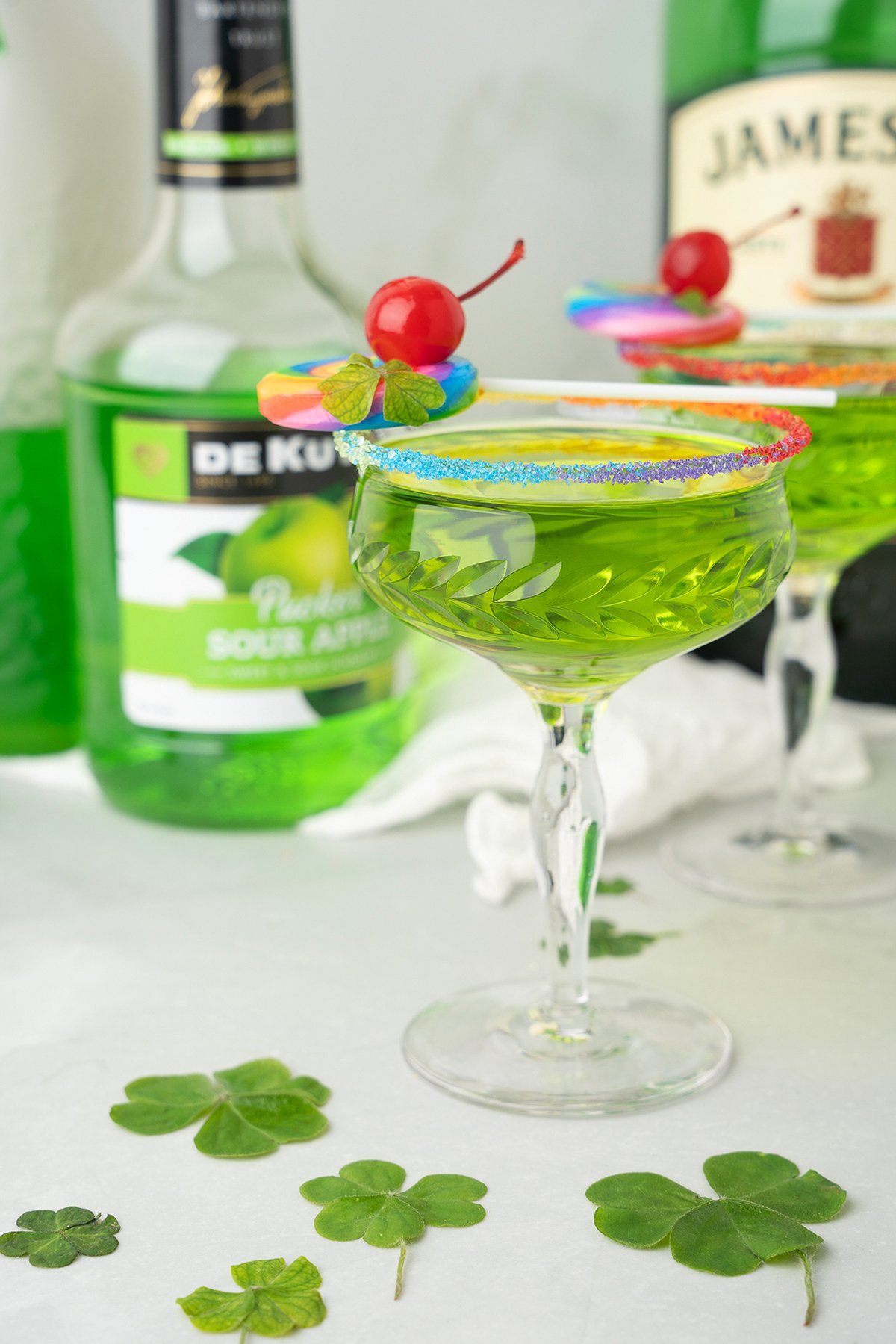 A green cocktail with sour apple schnapps in a rainbow sugar rimmed glass, topped with a cherry and rainbow garnish, surrounded by shamrock leaves.