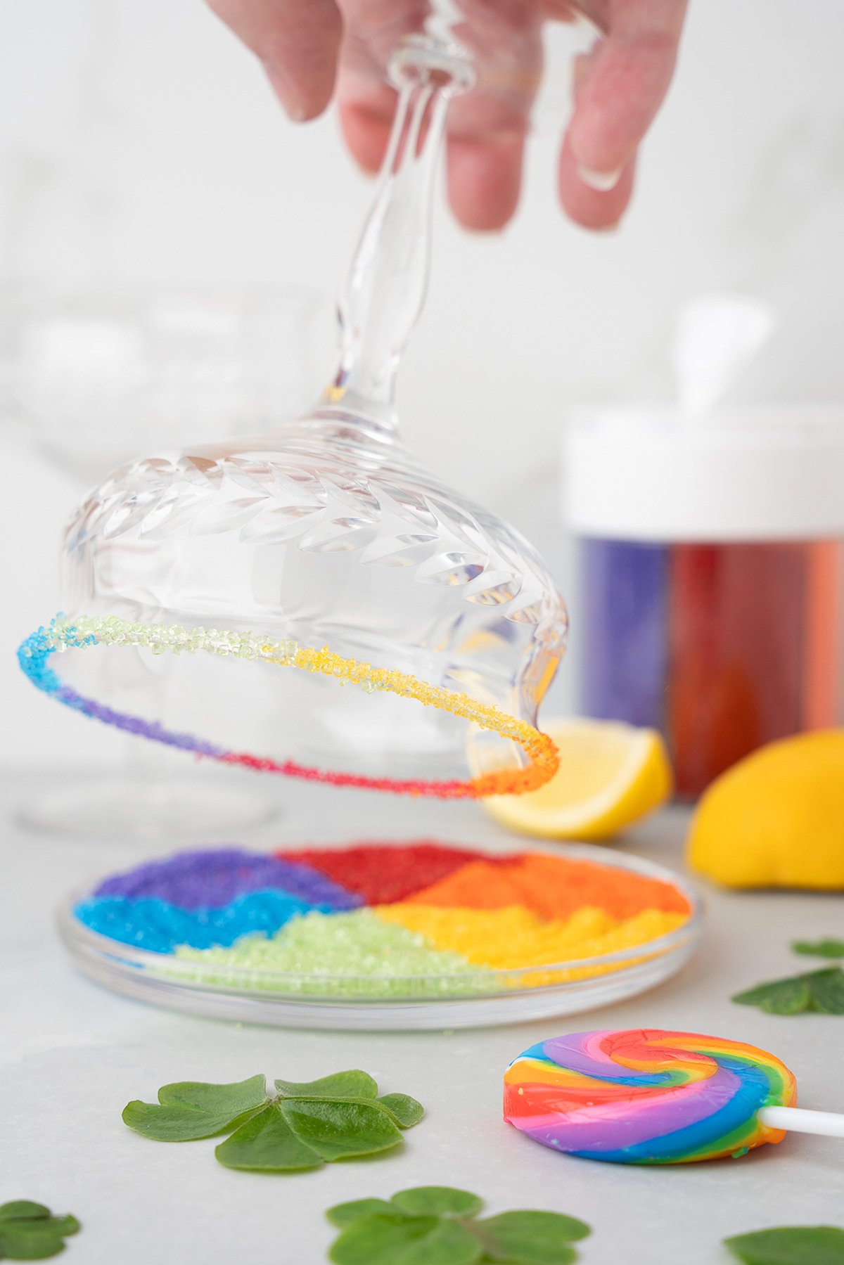 A glass is dipped into rainbow-colored sugar, perfect for a Rainbow Irish Whiskey Cocktail with melon liqueur, while a rainbow lollipop and lemons add festive flair—ideal for celebrating Pride Day.