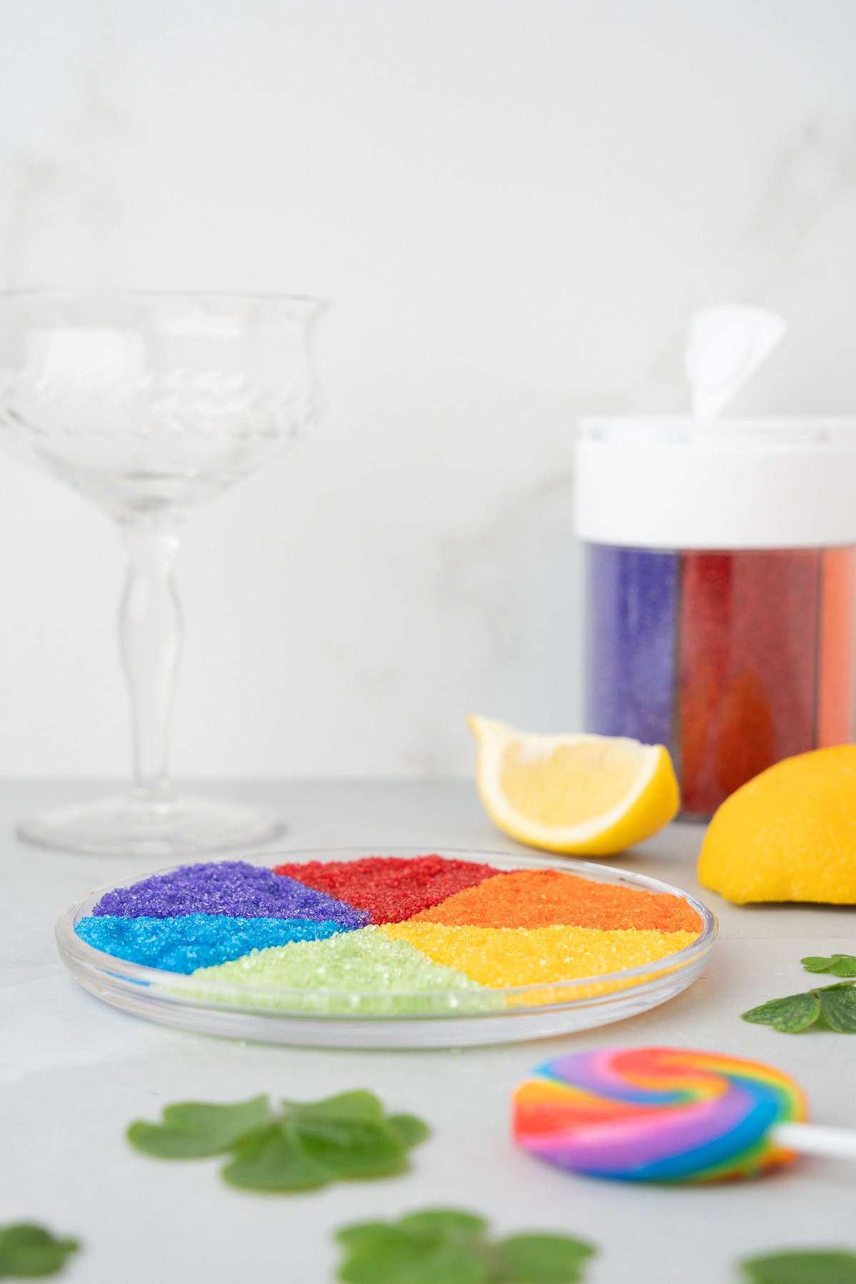 A plate with colorful sugar sprinkles, lemons, a rainbow lollipop, and an empty glass in the background—perfect inspiration for a Rainbow Irish Whiskey Cocktail on St. Patrick’s Day or Pride Day.