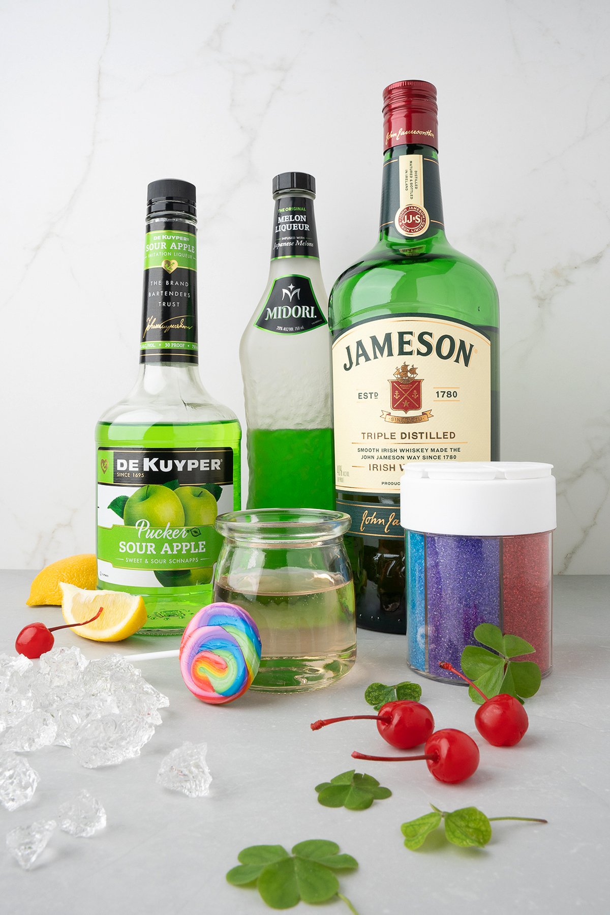 Bottles of liquor, cherries, ice, lemon, clover, and a rainbow swirl garnish on a white background—perfect elements for a Rainbow Irish Whiskey Cocktail with a festive rainbow sugar rim.