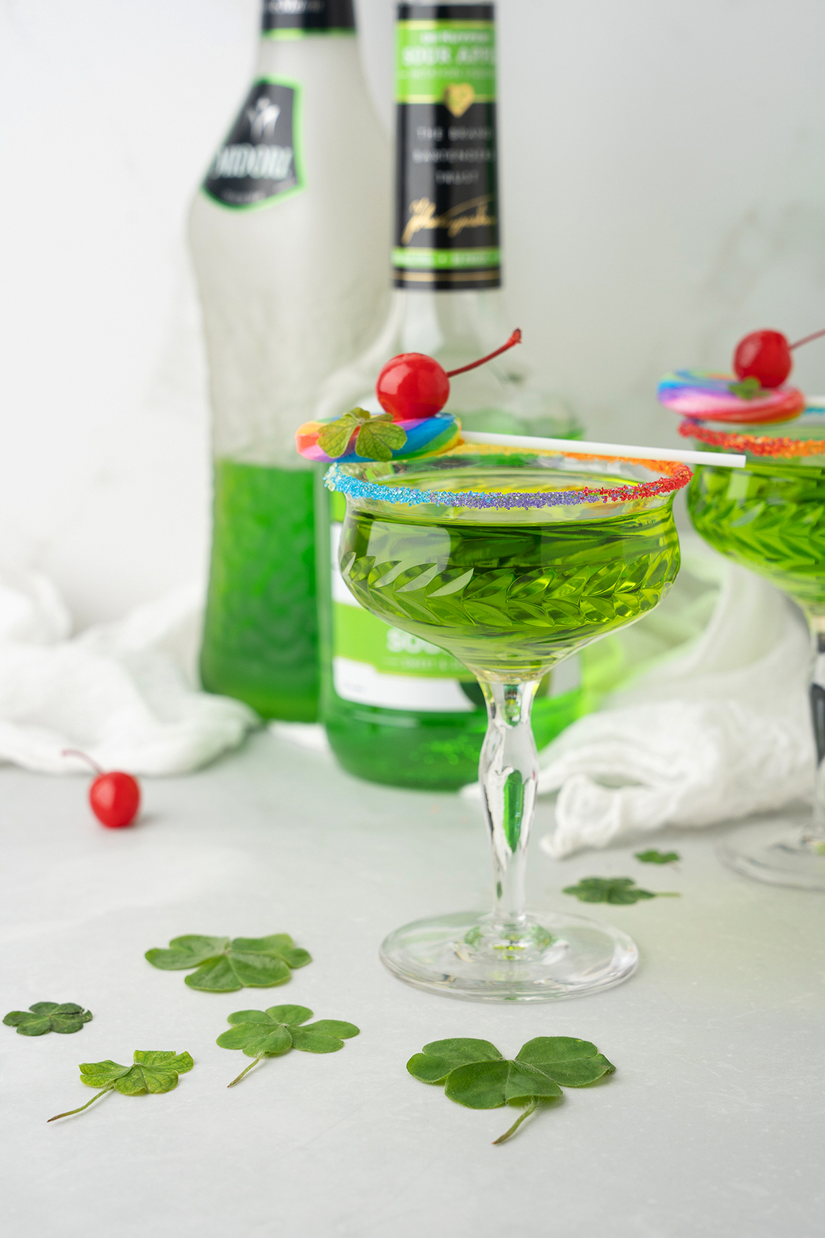 A vibrant Rainbow Irish Whiskey Cocktail featuring melon liqueur in a glass with a rainbow sugar rim, cherry, and clover—perfect for celebrating St. Patrick’s Day with colorful flair and green bottles in the background.