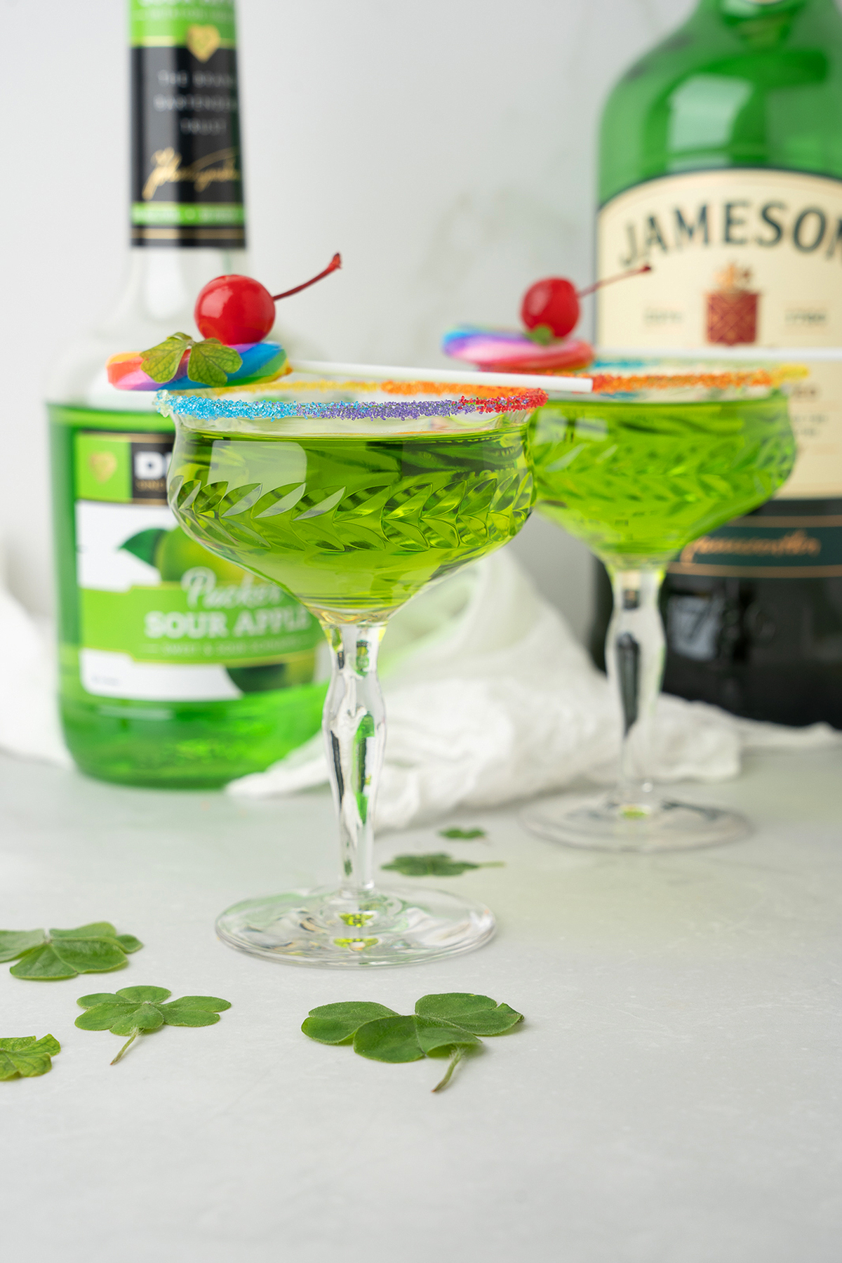 Two green Rainbow Irish Whiskey Cocktails in glasses with rainbow sugar rims, garnished with cherries and bottles in the background.