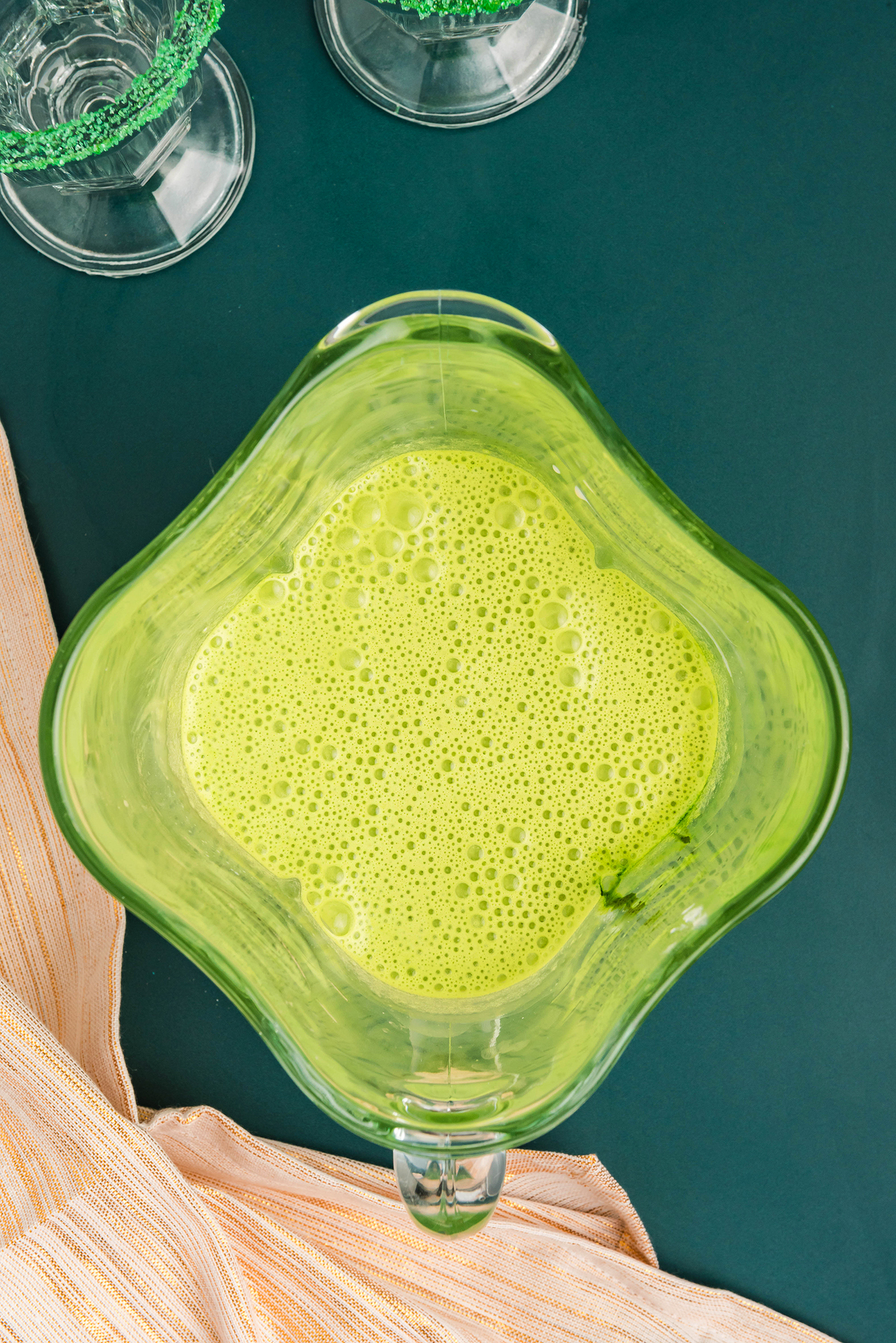 Top view of a blender jar filled with frothy, light green St. Patrick's shake on a dark green surface.
