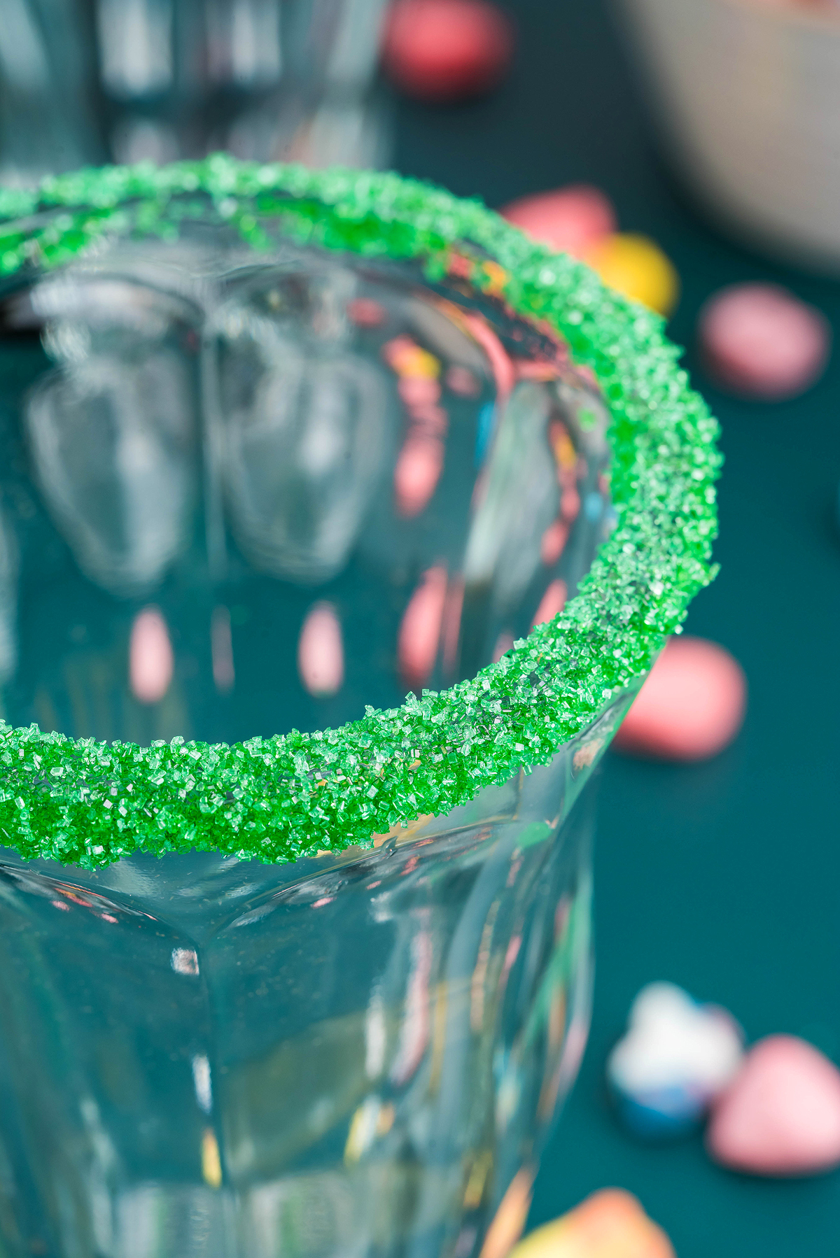 Glass with a rim coated in bright green sugar, capturing the festive vibe of a boozy shamrock shake, with blurred candies in the background.