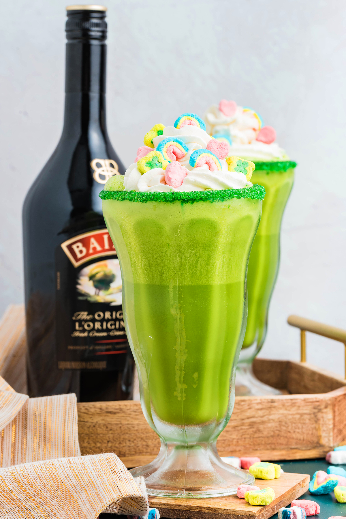 Green milkshakes with whipped cream and cereal toppings, next to a bottle of Baileys on a wooden tray—a festive Irish cream milkshake perfect for St. Patrick's Day celebrations.