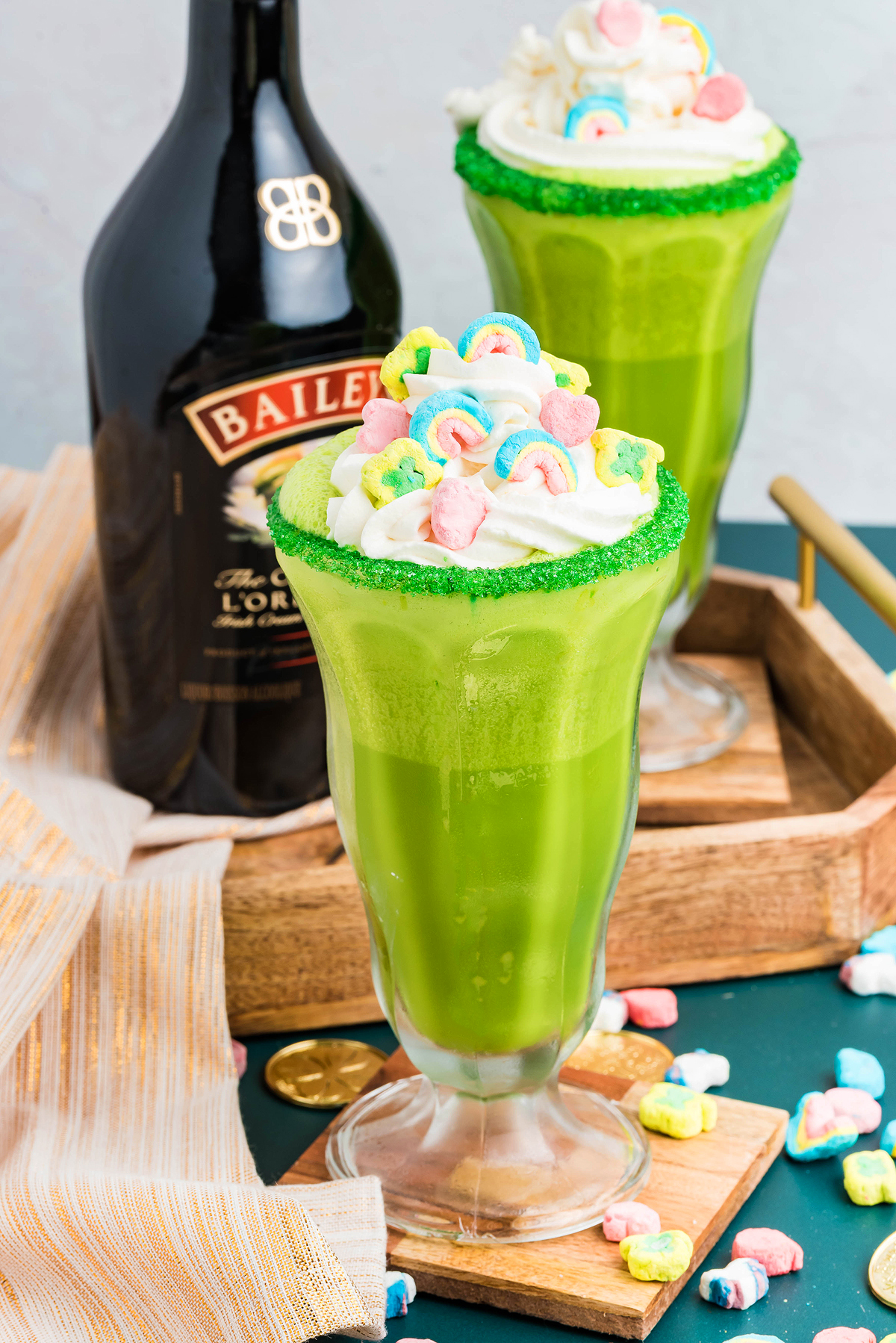 Two green drinks with whipped cream and colorful marshmallows, next to a Baileys bottle on a wooden tray—perfect for a festive boozy shamrock shake.