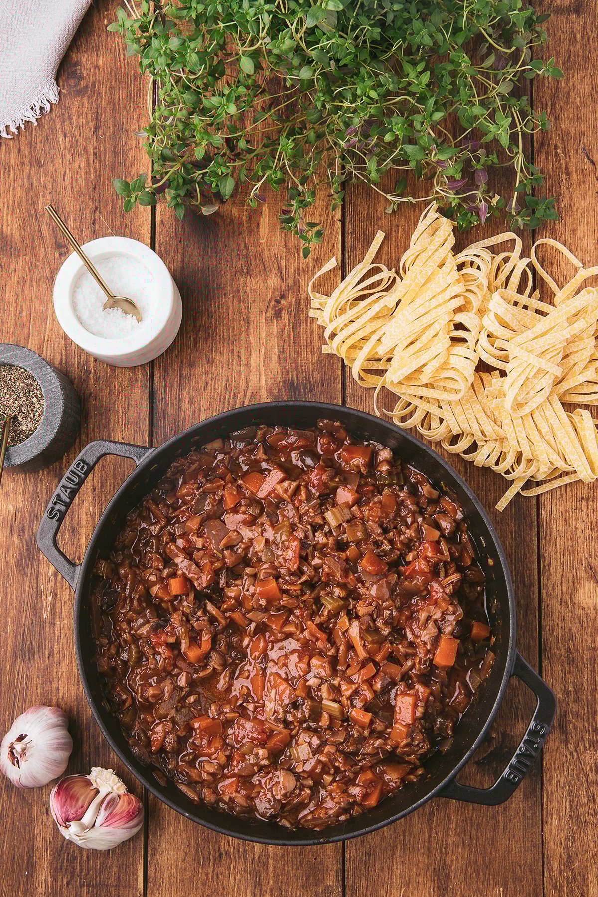 A pot of hearty homemade spaghetti sauce with mushrooms sits on a wooden table beside uncooked pasta, herbs, garlic, salt, and pepper.