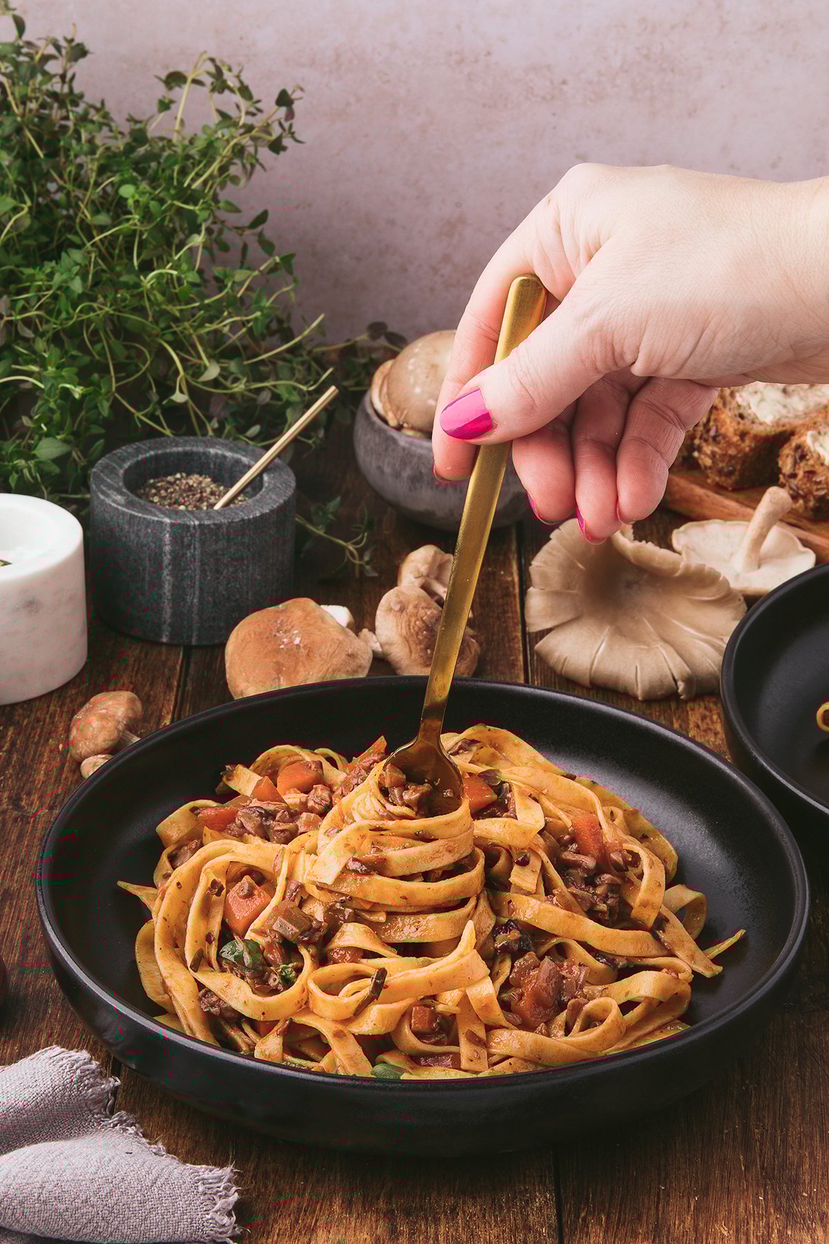 A hand twirls fettuccine pasta topped with Mushroom Ragu and fresh herbs in a black bowl on a rustic wooden table.