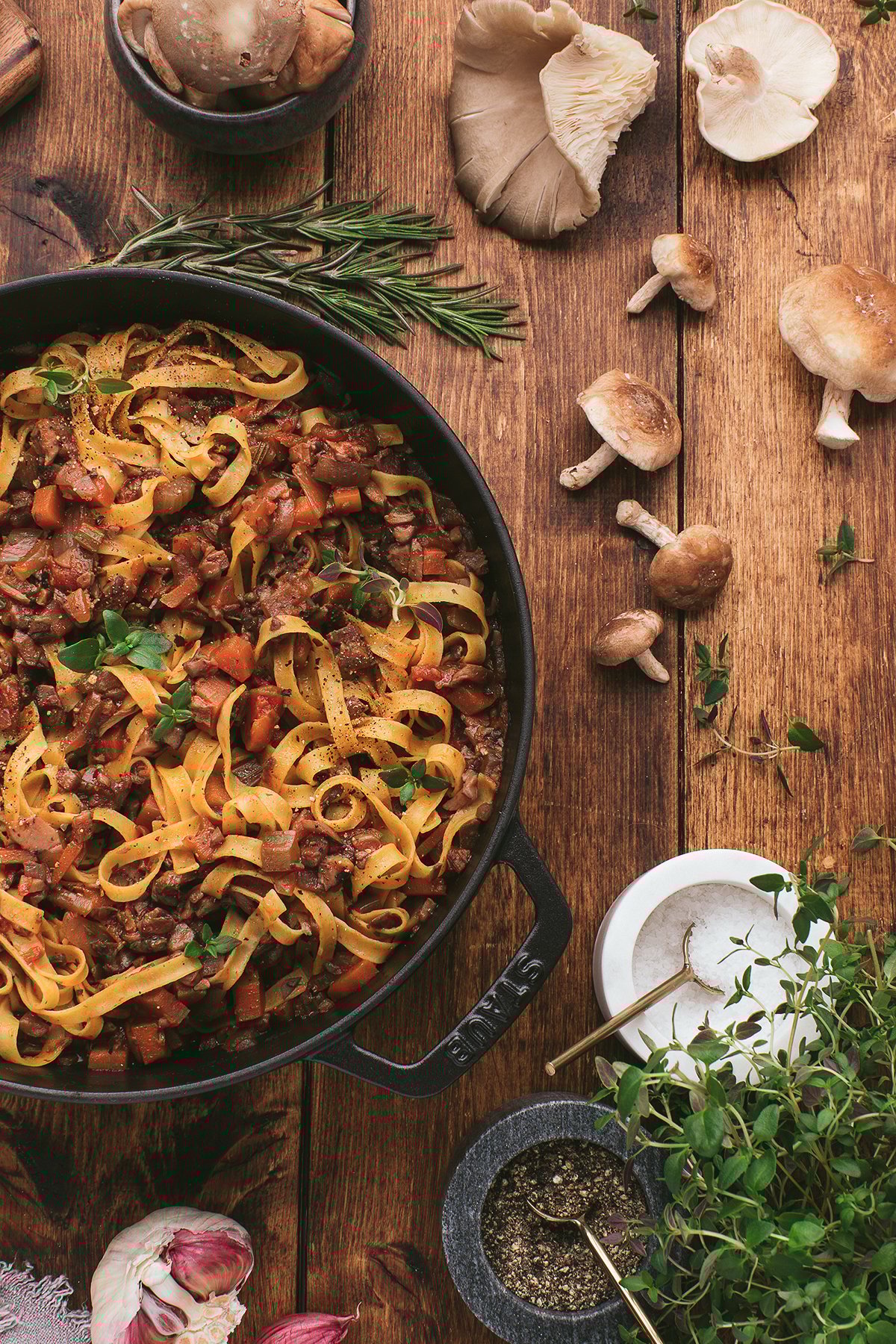 Tagliatelle pasta with a creamy vegan pasta sauce and mushrooms in a pan, surrounded by fresh herbs and whole mushrooms on a wooden table.