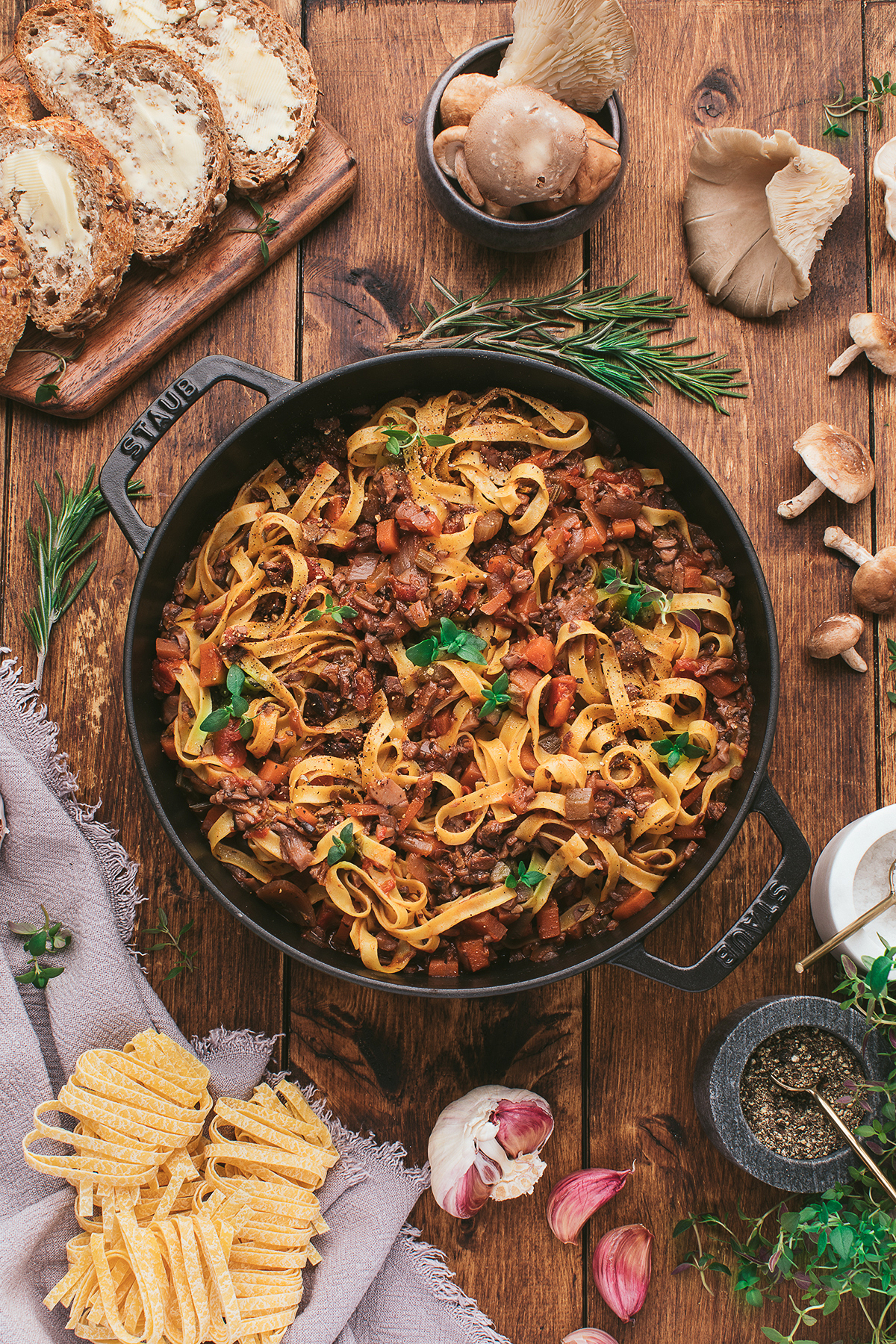 Cast-iron pot of tagliatelle pasta with a rich homemade spaghetti sauce with mushrooms, surrounded by bread, mushrooms, herbs, garlic, and uncooked pasta.