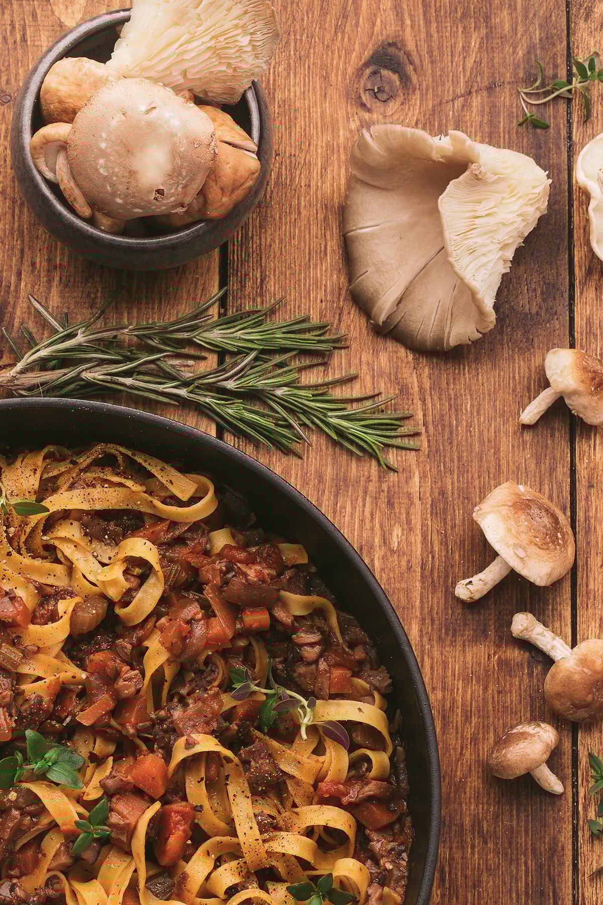 Tagliatelle pasta with a rich Vegan Pasta Sauce in a pan, surrounded by mushrooms and fresh rosemary on a wooden table.