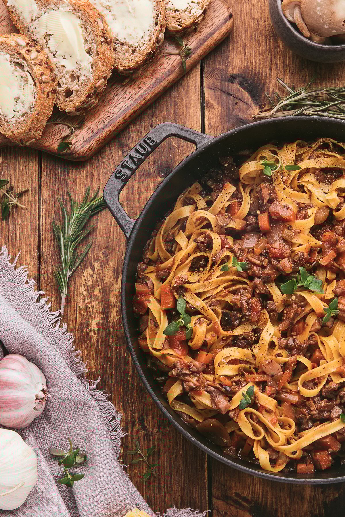 Tagliatelle pasta with homemade spaghetti sauce with mushrooms, served in a skillet alongside sliced bread, garlic, and fresh herbs on a wooden table.