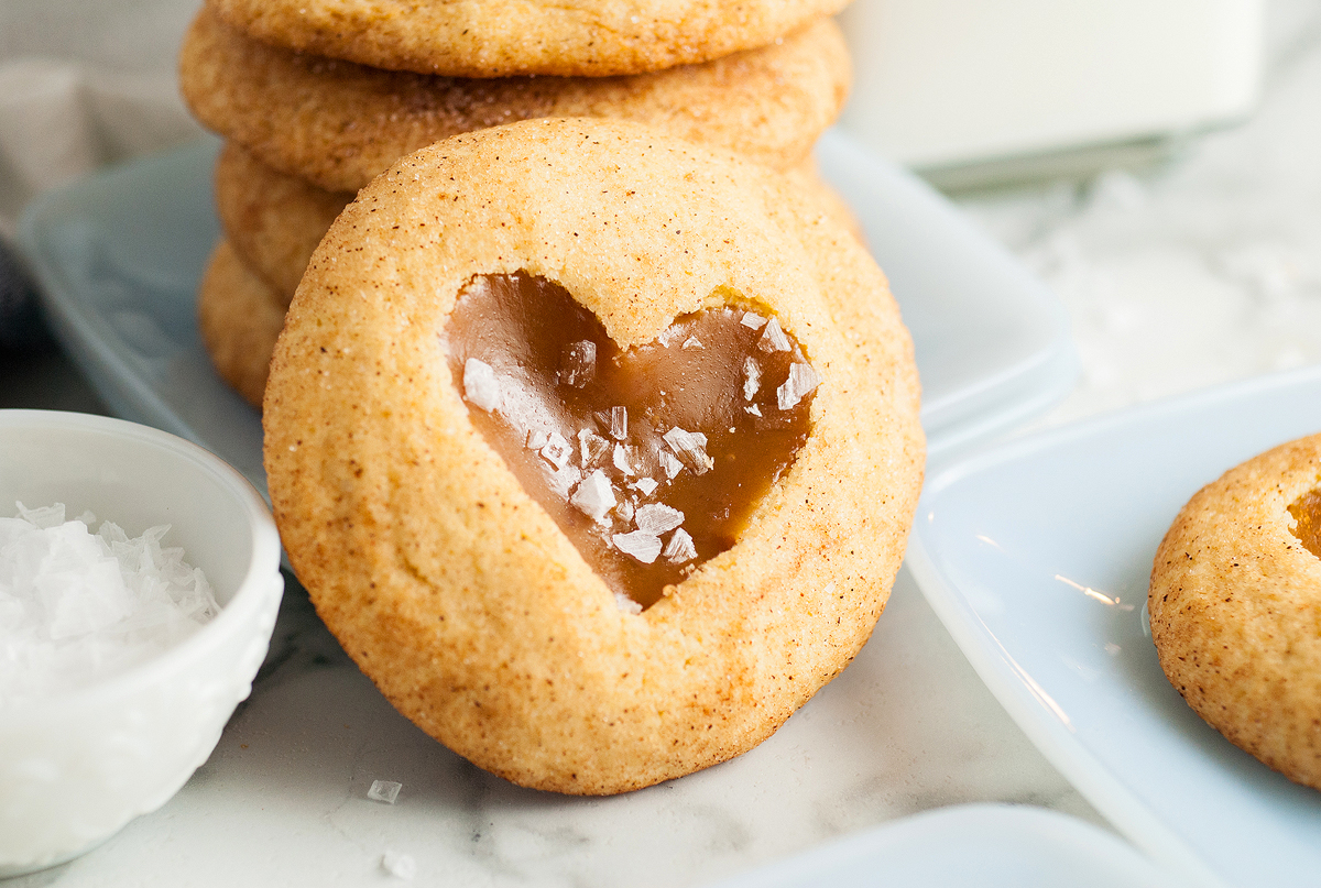 A close-up of a cookie with a heart-shaped caramel center topped with a few flakes of sea salt. The cookie is part of a small stack beside a bowl of salt and a glass of milk, all situated on a light-colored surface.