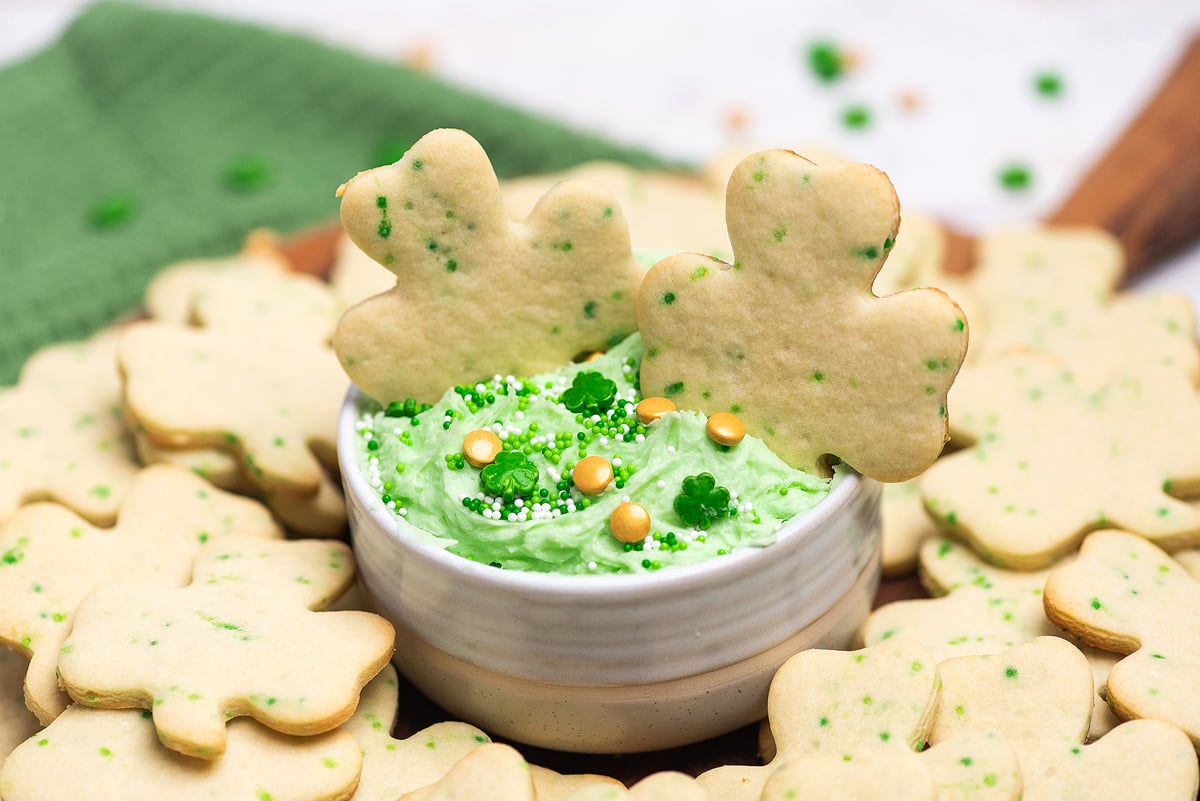 Shamrock sugar cookies with green sprinkles and gold accents make perfect treats for St. Patty's party ideas—festive like Dunkaroos, they're topped with green frosting and white decorations in a bowl.