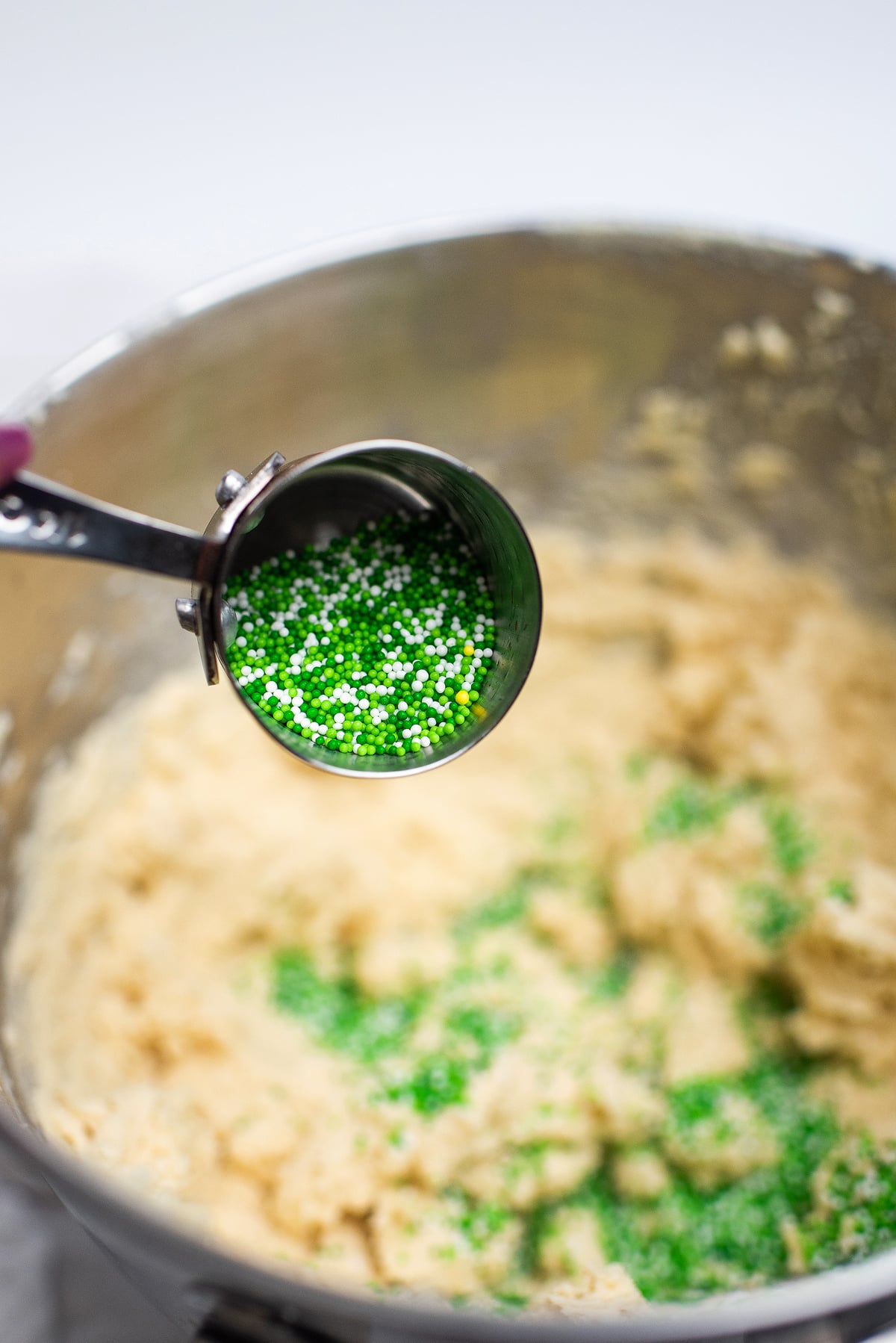A measuring cup pours green and white sprinkles into a bowl of cookie dough, perfect for making festive shamrock sugar cookies for your St. Patrick's Day cookie recipe.