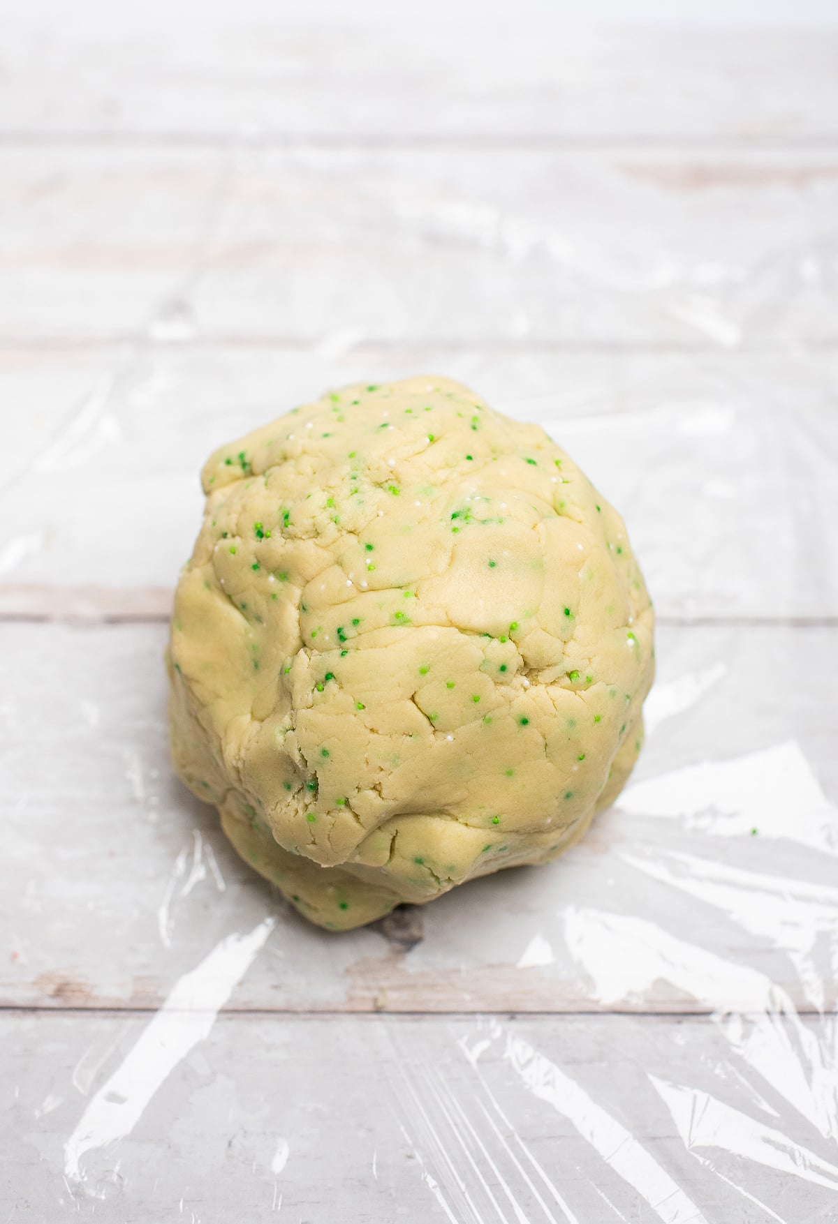 A ball of cookie dough with green sprinkles, perfect for St. Patrick's Cookies and Buttercream Dip, sits on clear plastic wrap atop a white wooden surface—an ideal treat for St. Patty's party ideas!.