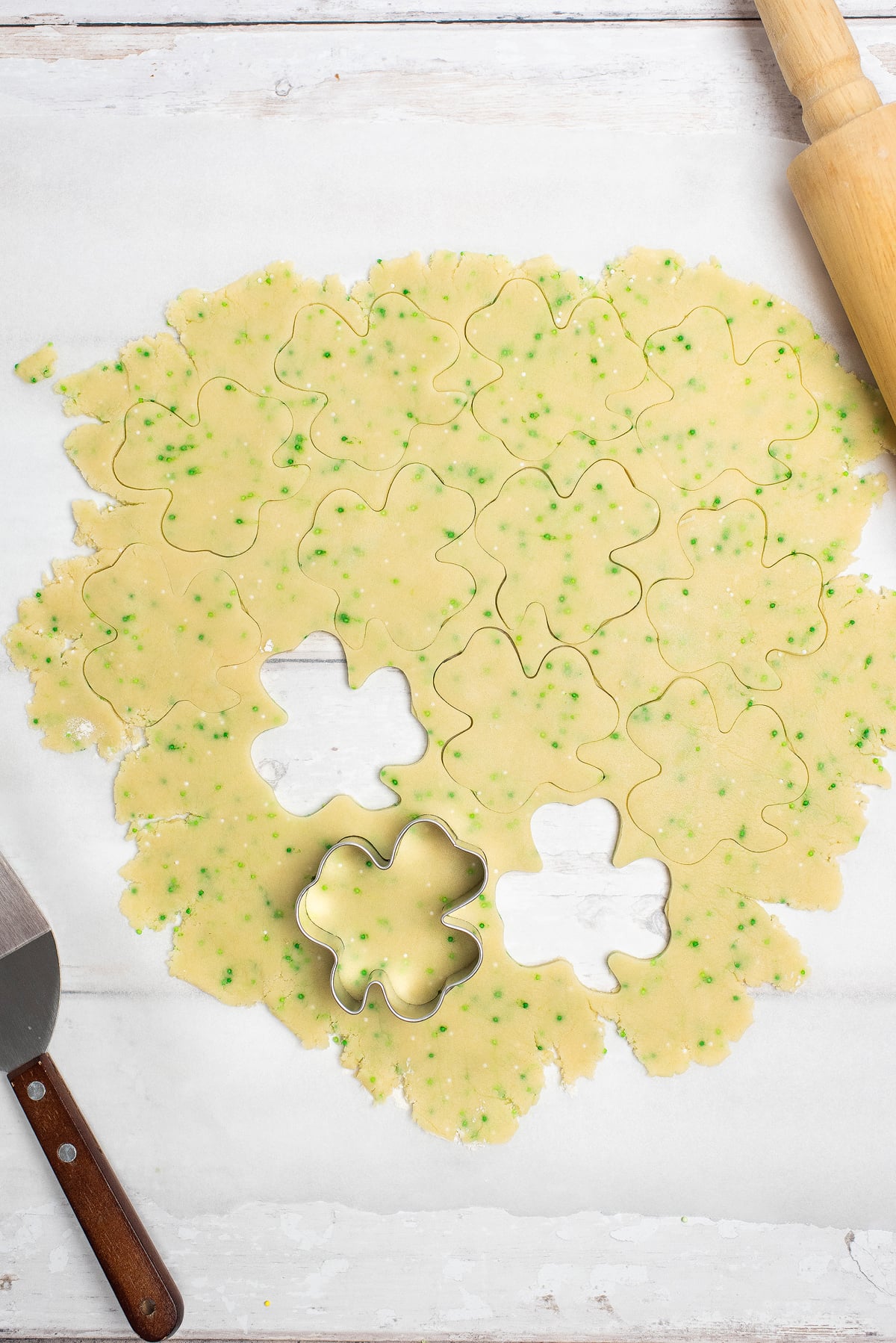 Shamrock sugar cookies are cut from dough and topped with green sprinkles on a white surface, with a rolling pin nearby—perfect for festive St. Patrick's Cookies and Buttercream Dip.