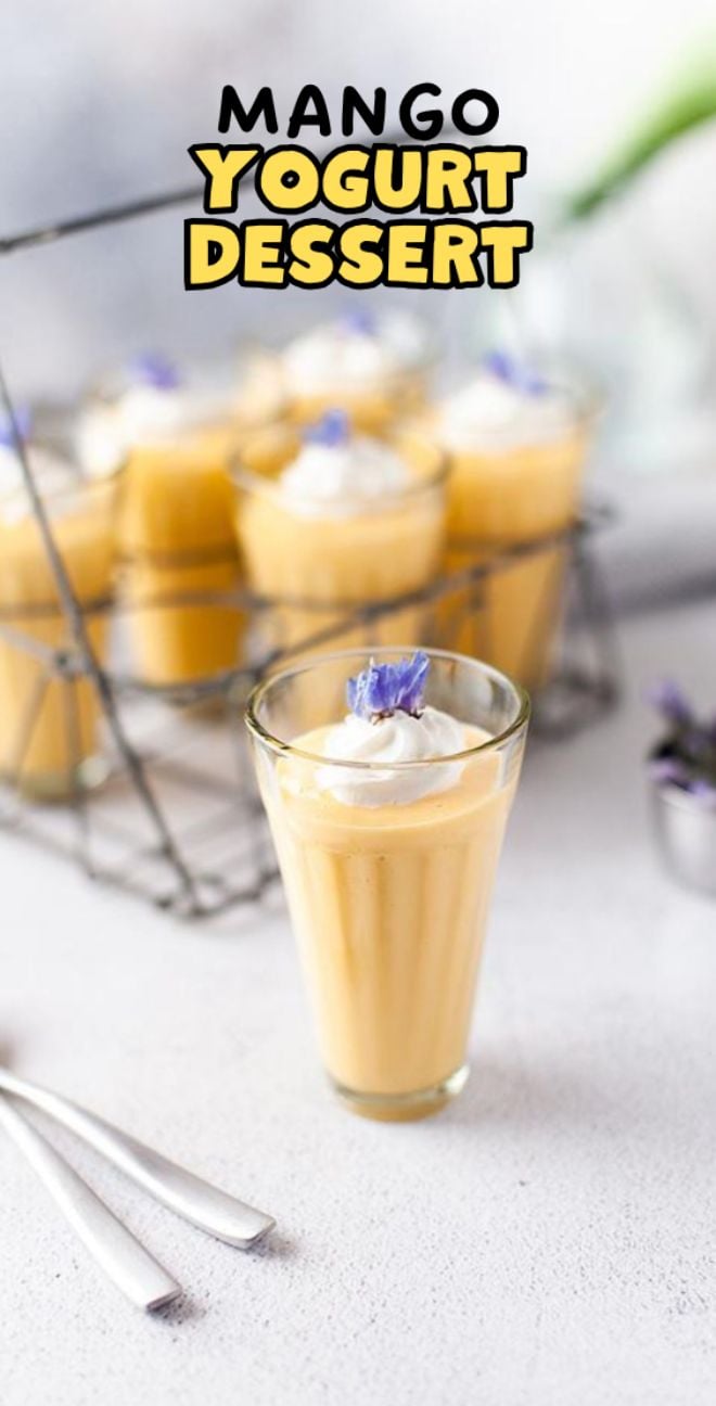 A glass of mango yogurt dessert topped with cream and a purple flower, with more desserts in the background—an inviting treat inspired by a healthy mango mousse recipe.