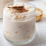 A glass of Mexican rice pudding, or Arroz Con Leche, is topped with cinnamon sticks and a sprinkle of cinnamon, resting on a white tile surface. Nearby lies a spoon dusted with cinnamon and a golden measuring cup.