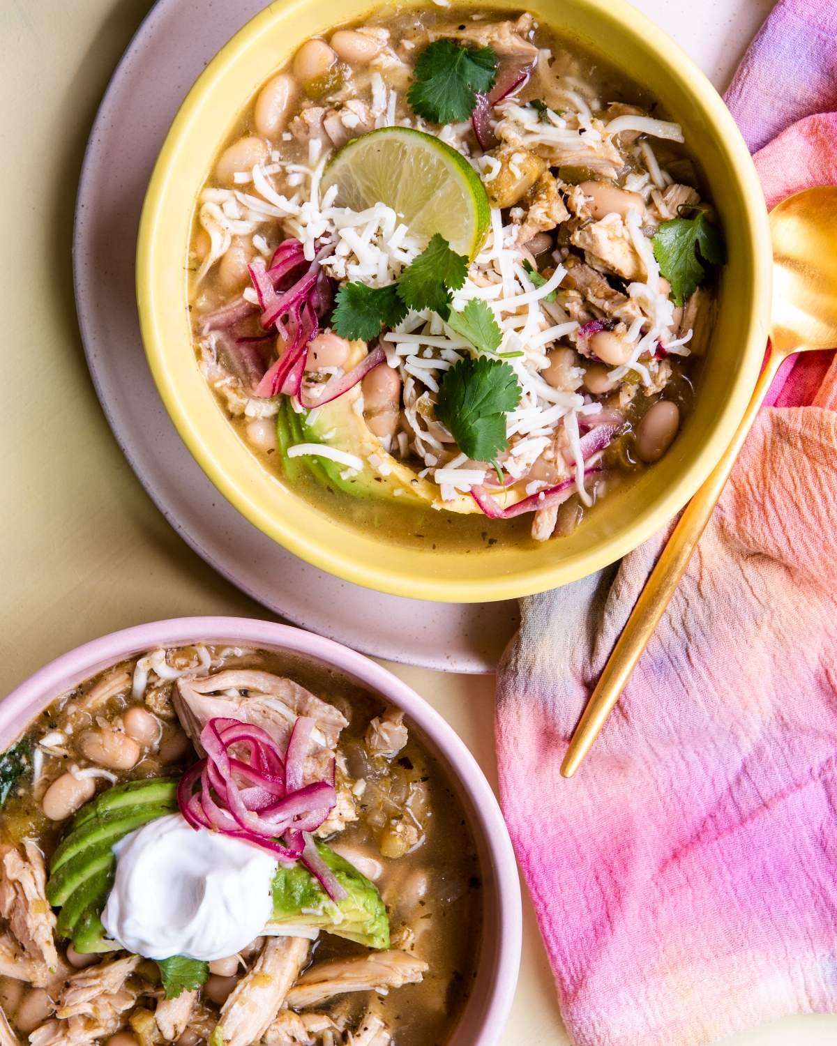 Two bowls of Mexican inspired white chili topped with cheese, cilantro, avocado, pickled onions, lime, and sour cream.