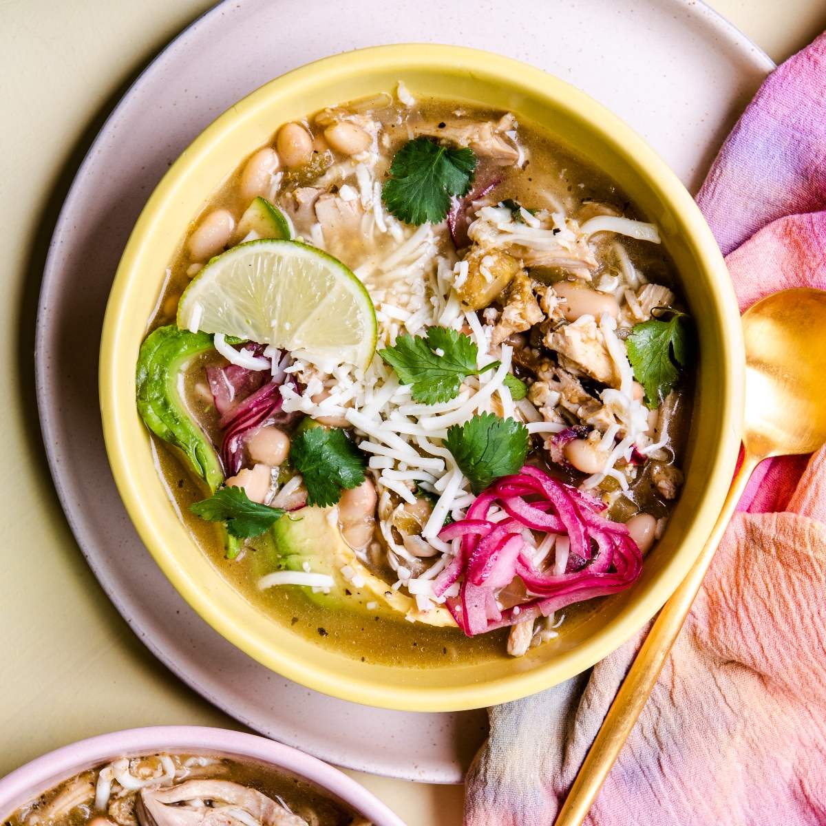 Bowl of Mexican inspired white chicken chili topped with cheese, lime, avocado, cilantro, and pickled onions, served with a gold spoon.