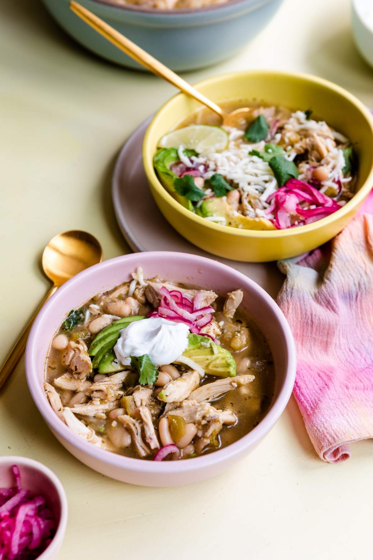 Two bowls of Mexican inspired chili verde chicken soup topped with sour cream, avocado, cilantro, pickled onions, and rice.