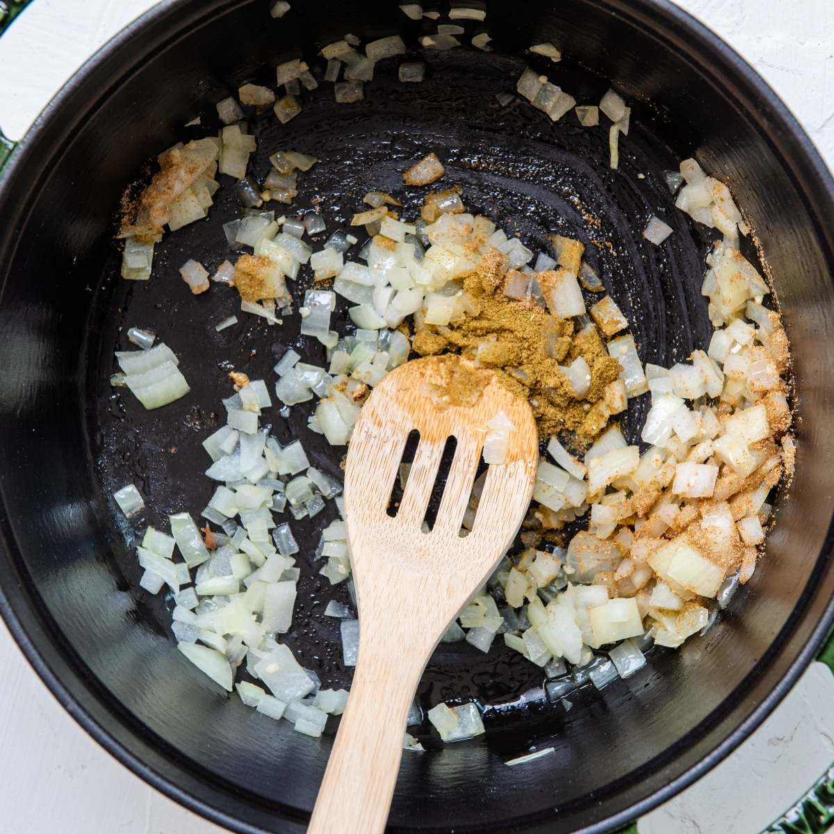 Chopped onions and spices are being sautéed in a black pan with a wooden slotted spoon, creating the flavorful base for a delicious White Bean Green Chile Chicken Soup.