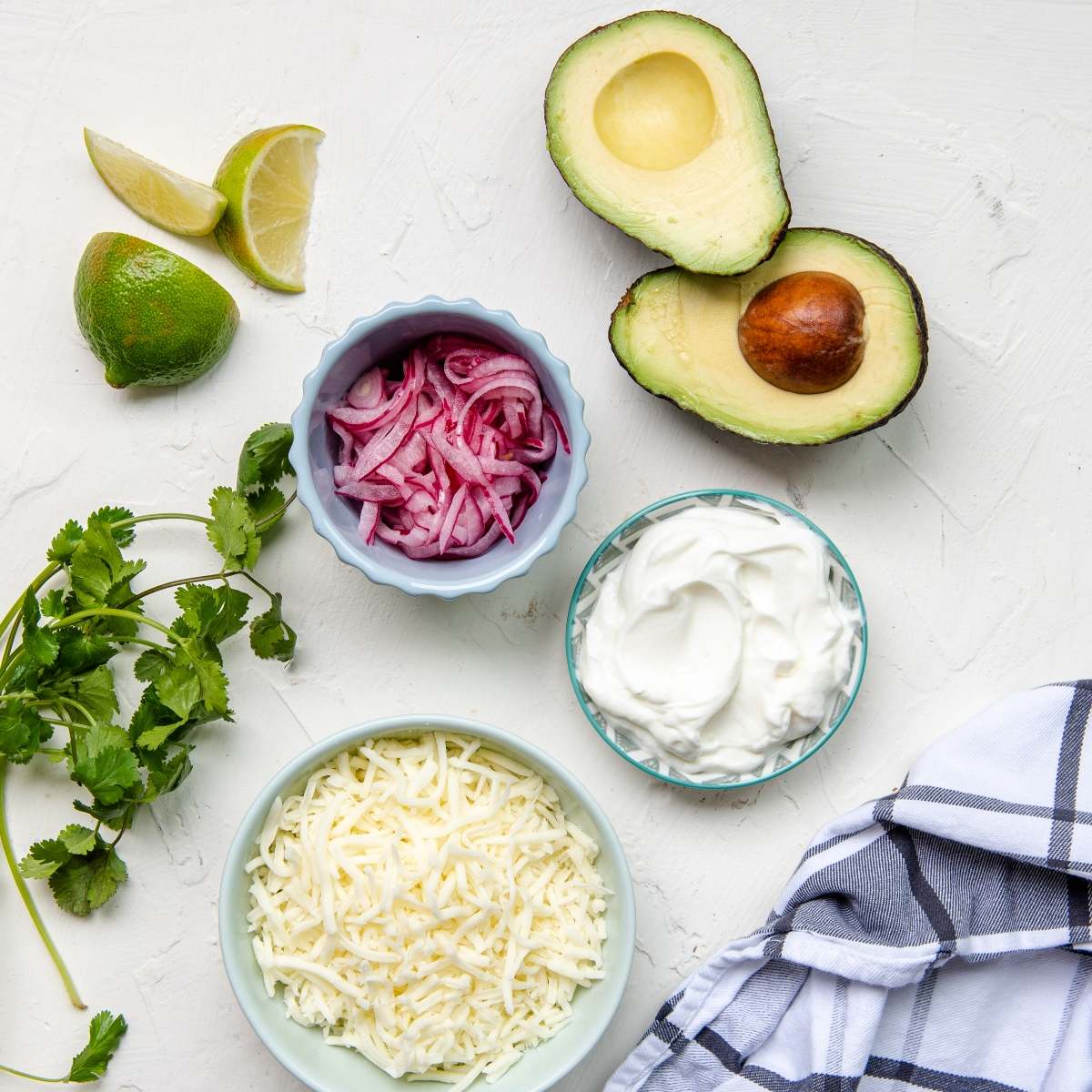 Sliced avocado, lime, pickled onions, shredded cheese, sour cream, and cilantro on a white surface—perfect toppings for a Mexican inspired green chili soup.