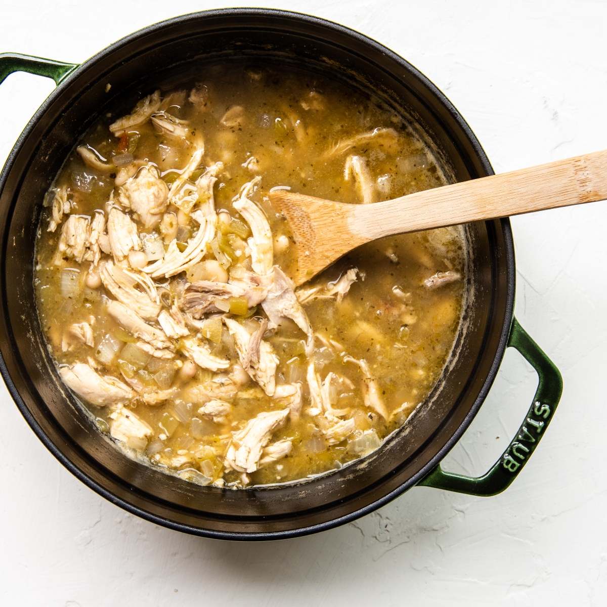 Mexican inspired White Bean Green Chile Chicken Soup with shredded chicken and beans in a green pot, stirred with a wooden spoon on a white background.